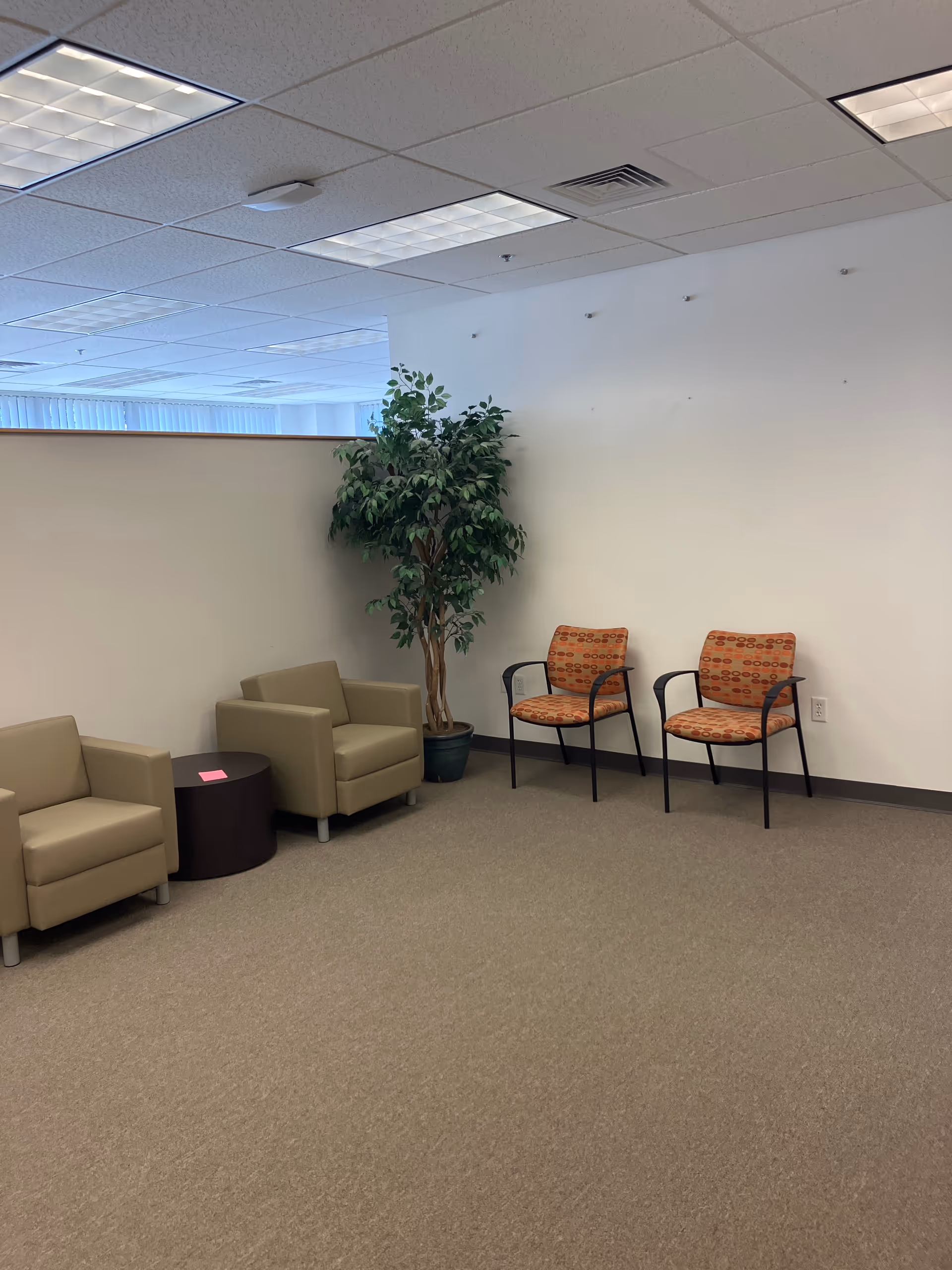 A waiting area with beige lounge chairs, orange patterned chairs, and a tall plant.