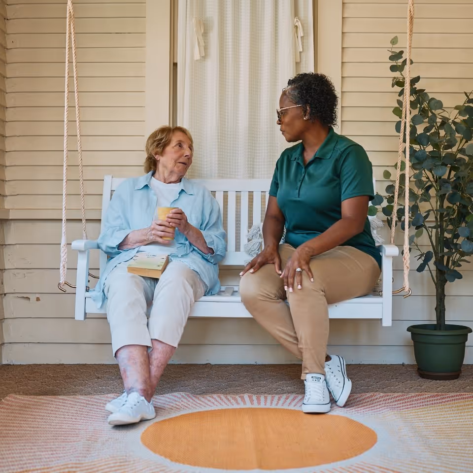 Two women sit on a white porch swing, chatting outside a home.