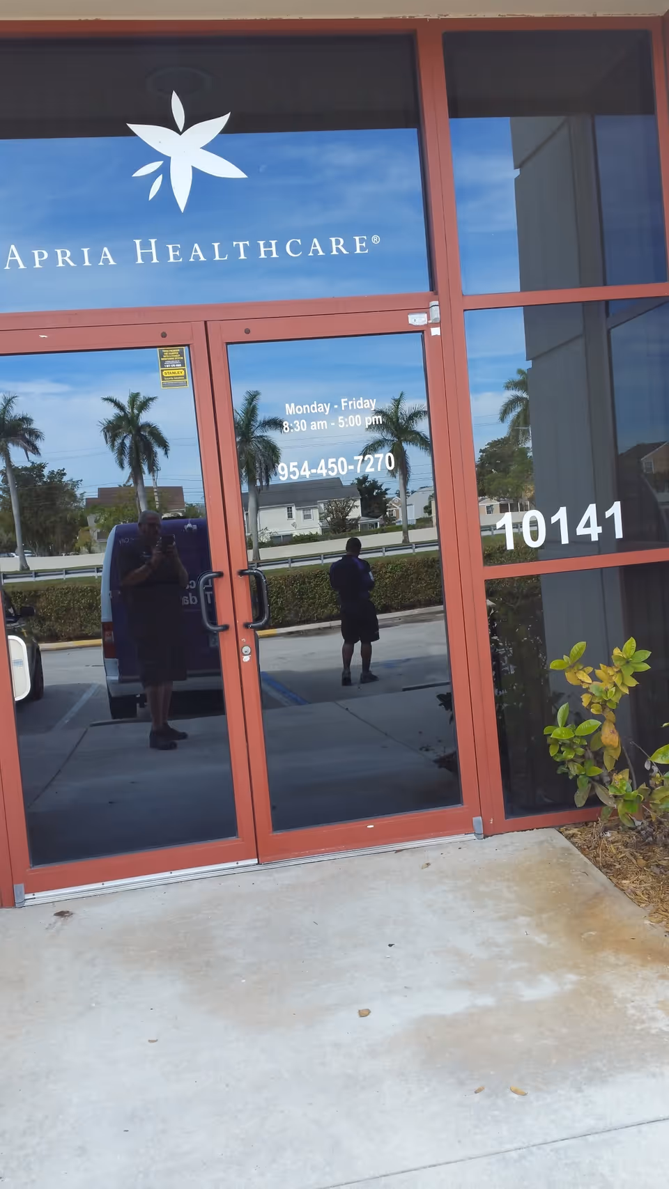 Apria Healthcare glass entrance with red frame; two people reflected outside.