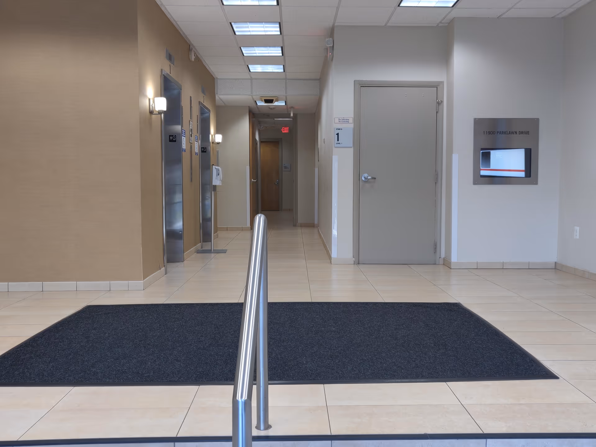 Bright interior hallway with elevators, a gray door, wall display panel, and metal handrail.
