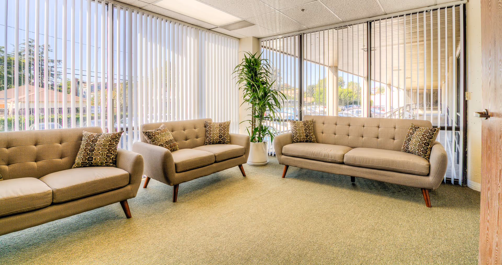 Beige lounge area with three tufted sofas, a tall plant, and large windows with blinds.