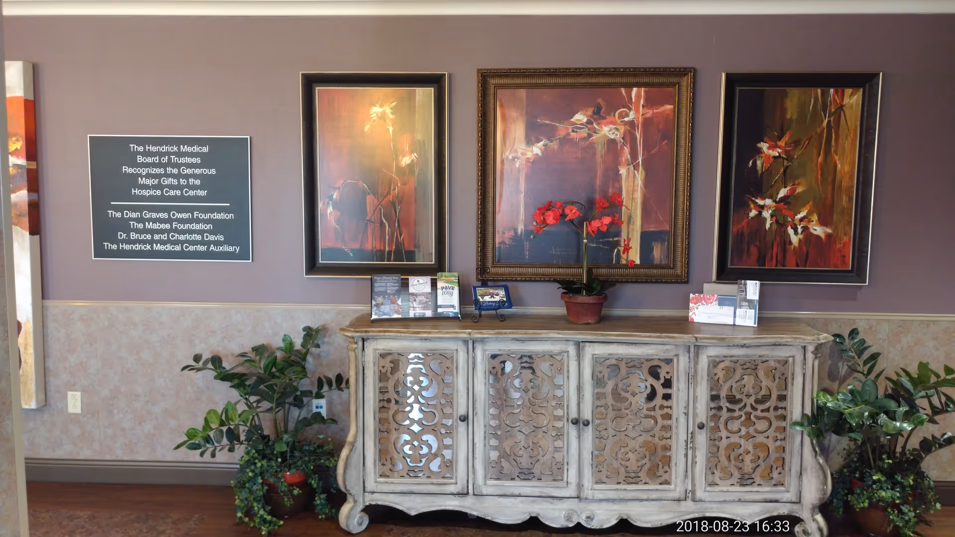 Decorative antique cabinet with floral artworks, potted plants, and a plaque on a purple wall.