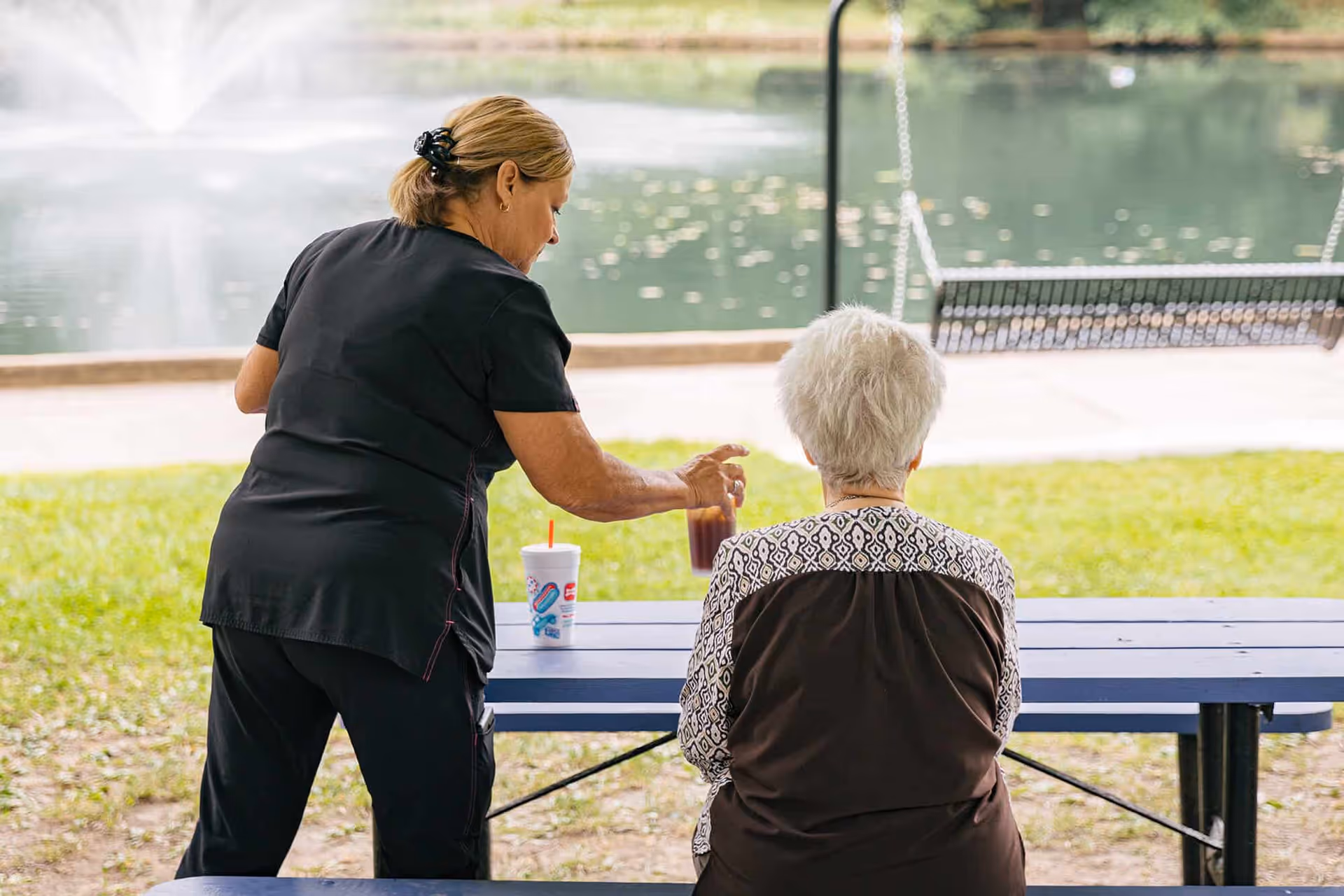A caregiver assists an elderly resident at a blue picnic table beside a park pond.