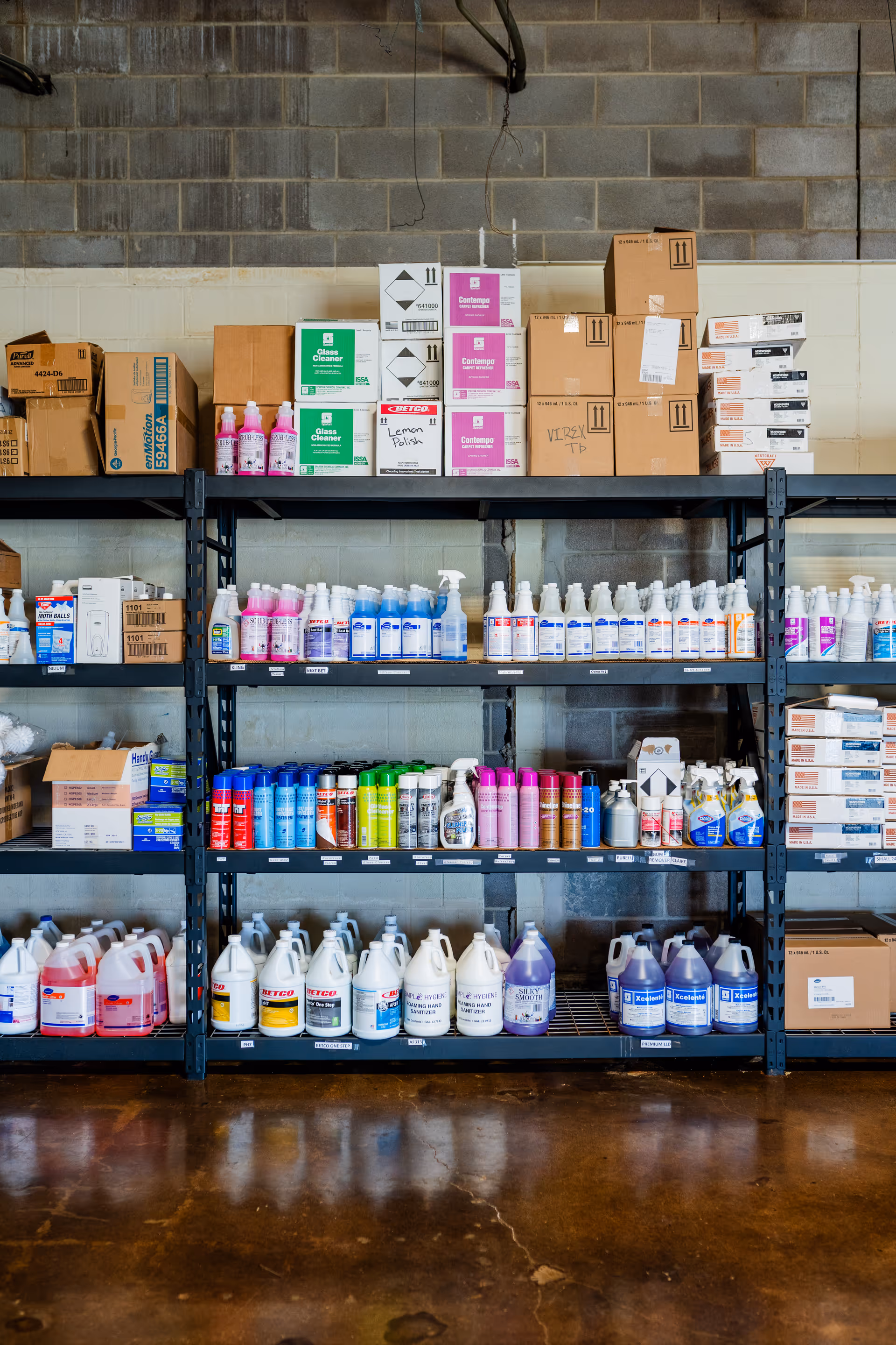 Industrial storage shelves filled with cleaning products and cardboard boxes against a concrete wall.