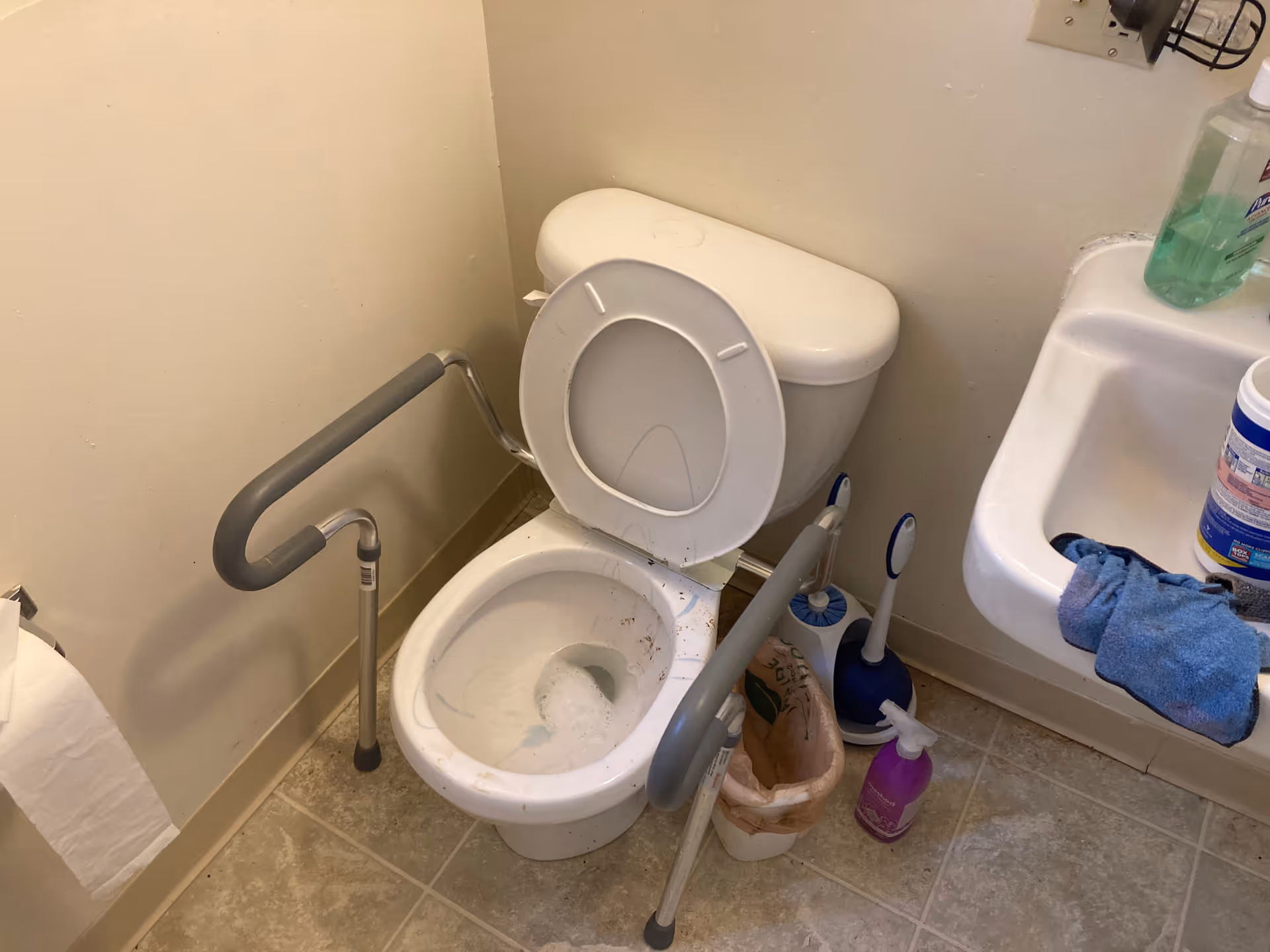 Accessible bathroom with a toilet, grab bars, and sink; cleaning supplies visible.