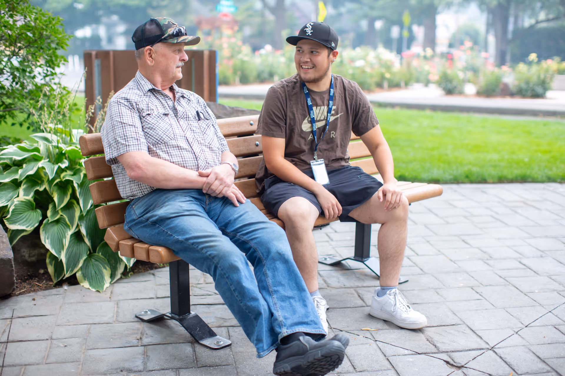 Two men sit on a wooden bench in a sunny outdoor park, chatting.