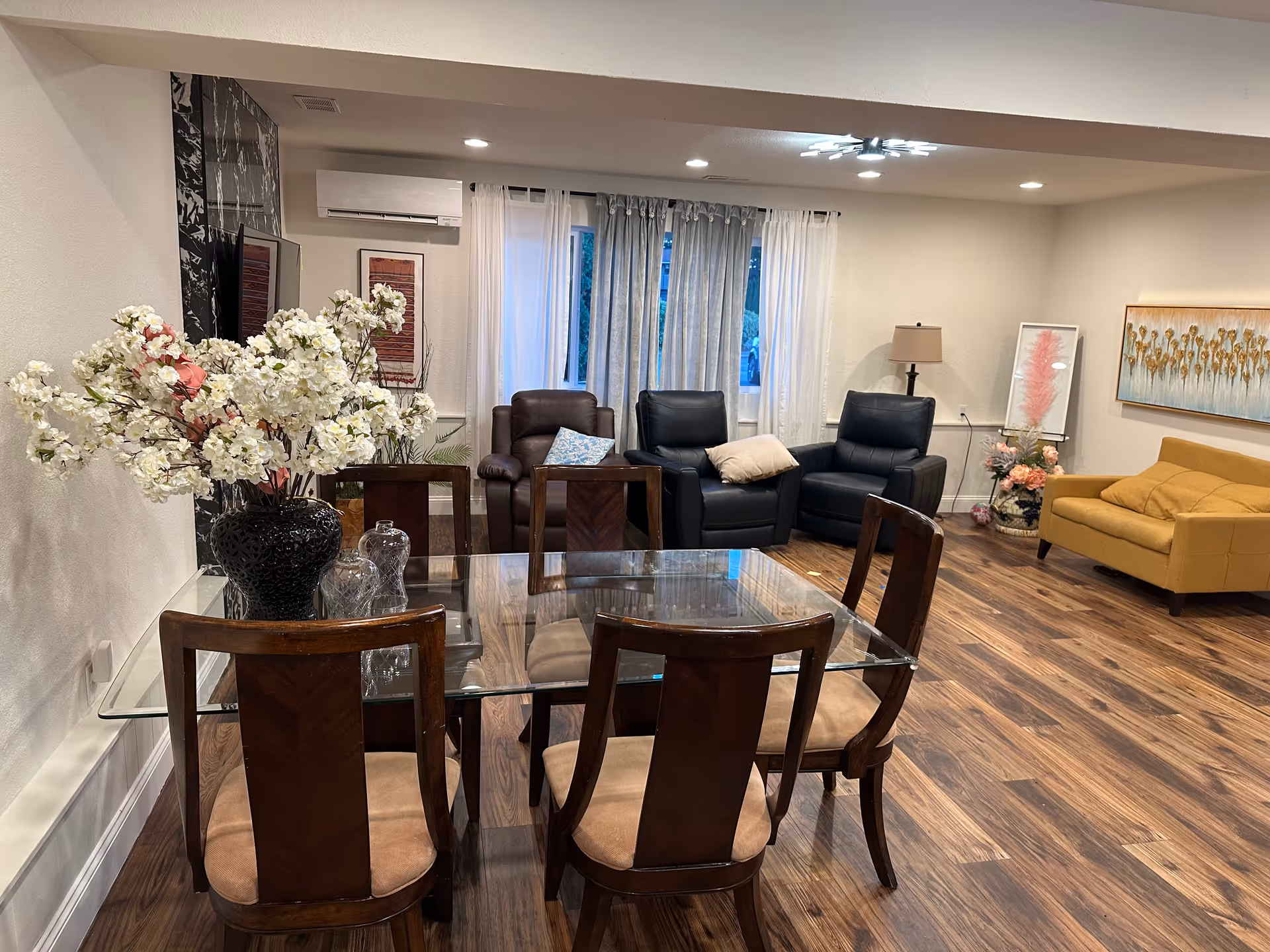 Well-lit senior living common area with glass dining table and plush seating.
