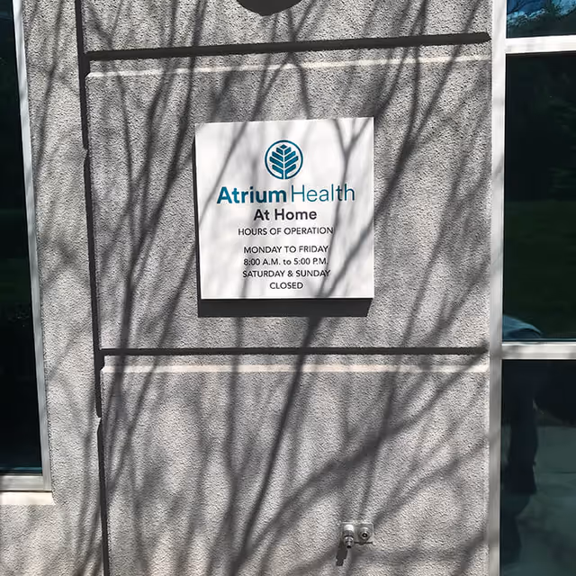 Exterior wall sign reads Atrium Health At Home with hours posted, tree shadows.