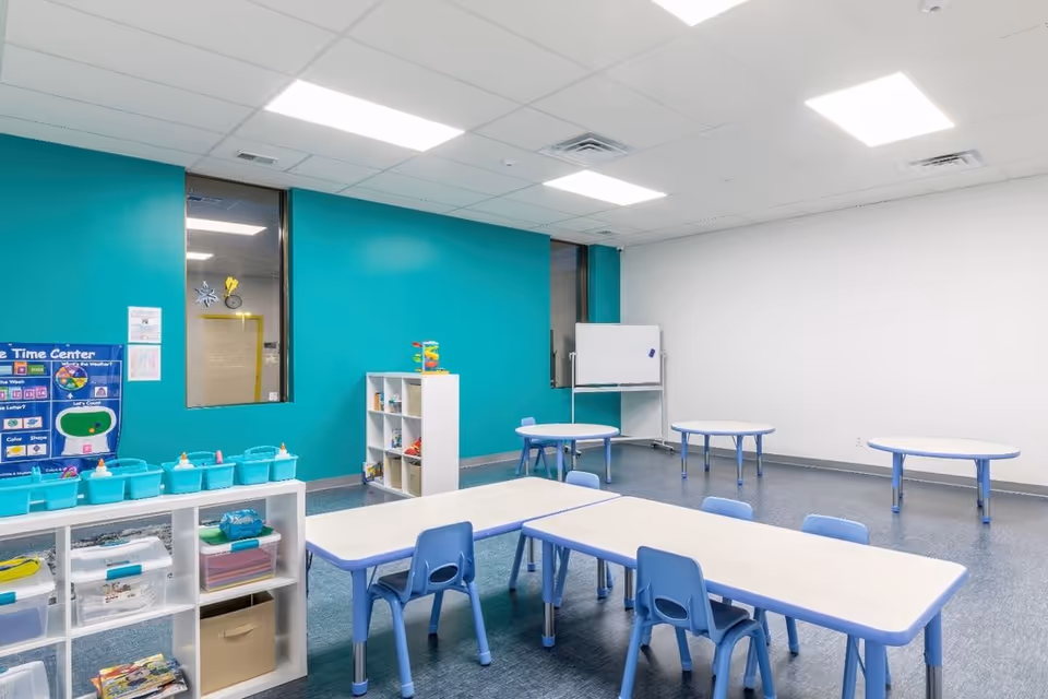 Bright therapy classroom with blue chairs, tables, teal accent wall, and shelves.