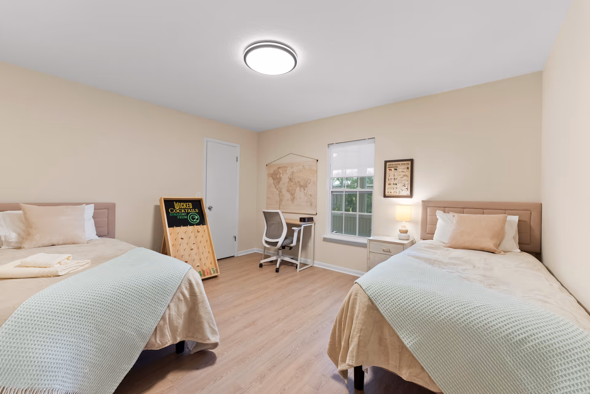 Two twin beds in a beige, well-lit bedroom with desk, map, and lamps.