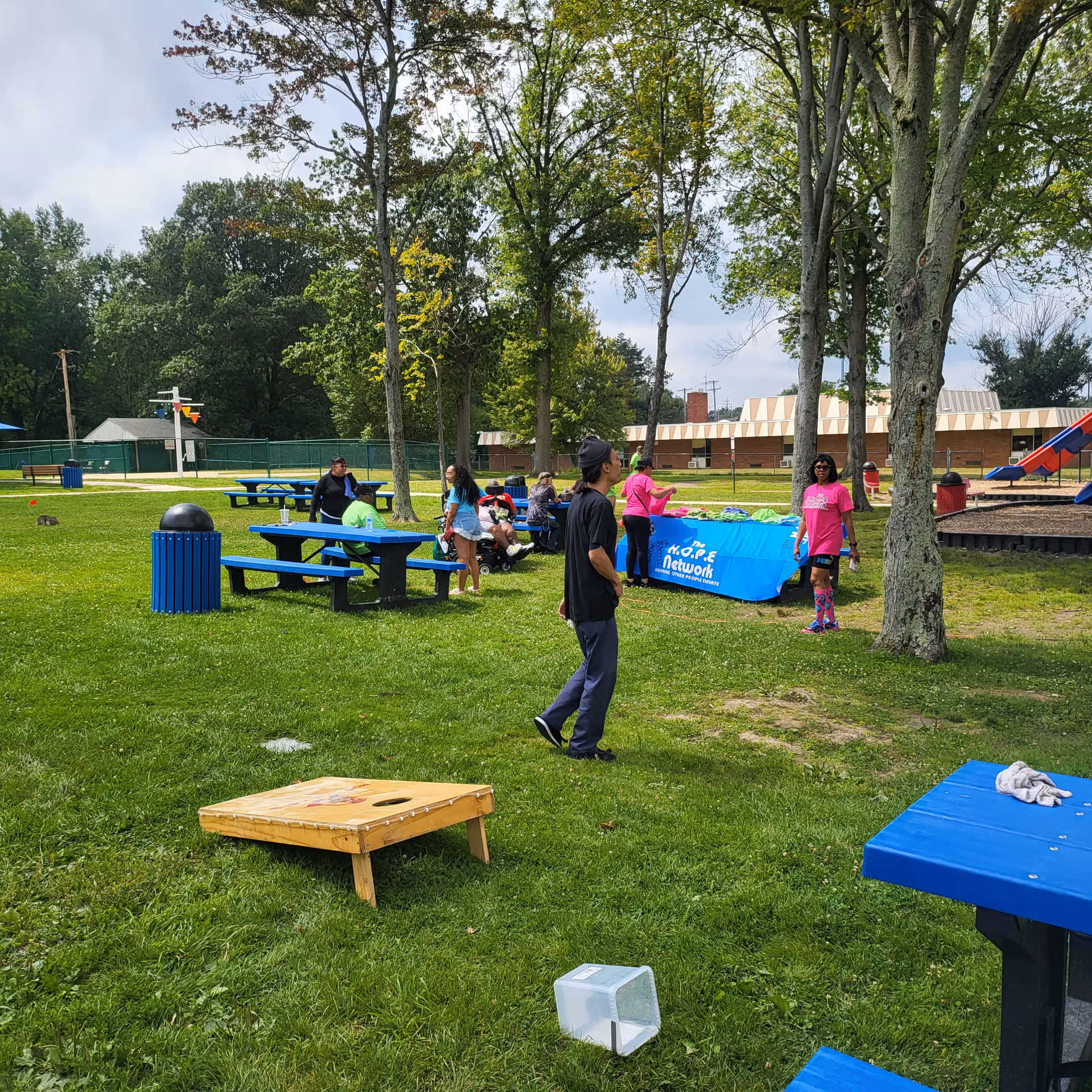 People gather outdoors at a park-like area with picnic tables and trees.