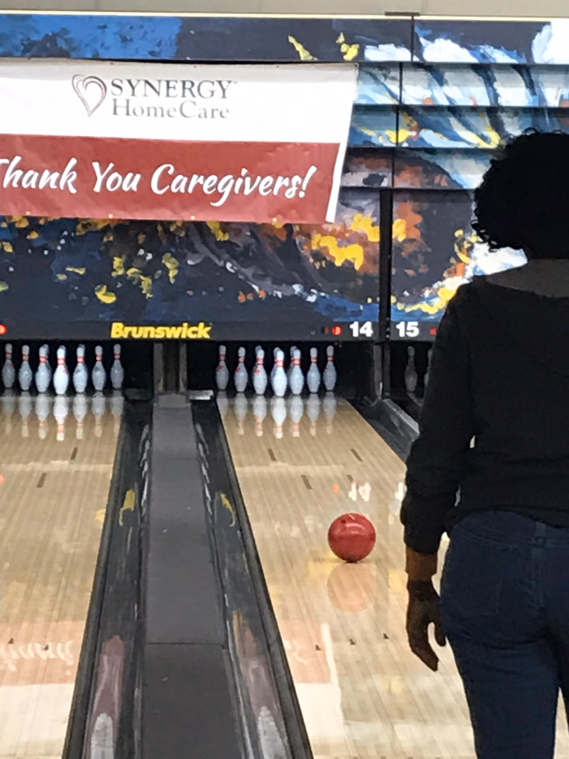 A person stands at a bowling lane beside pins and a SYNERGY HomeCare banner.