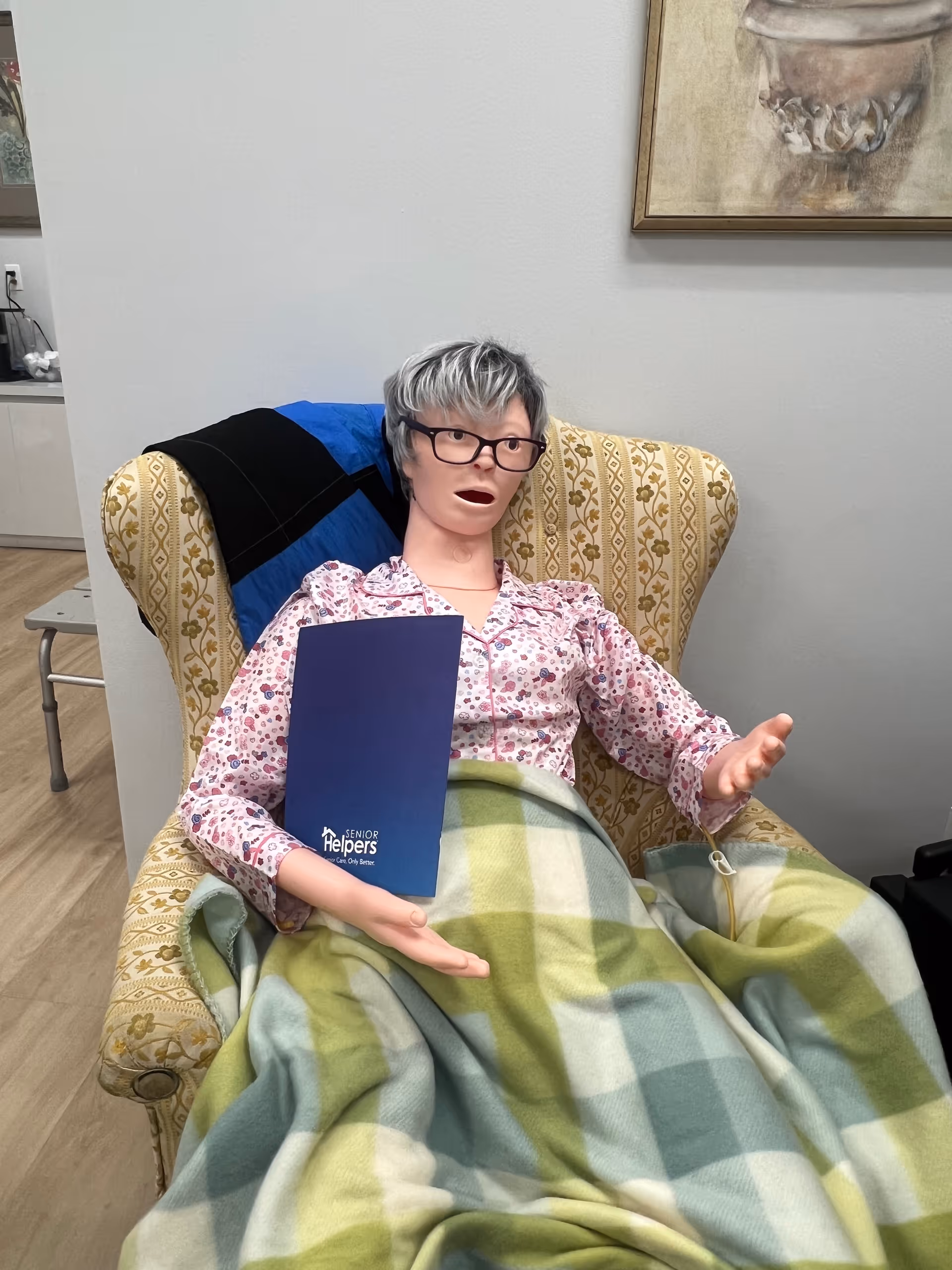 Life-sized mannequin in a patterned armchair, holding a blue Senior Helpers folder, blanket nearby.