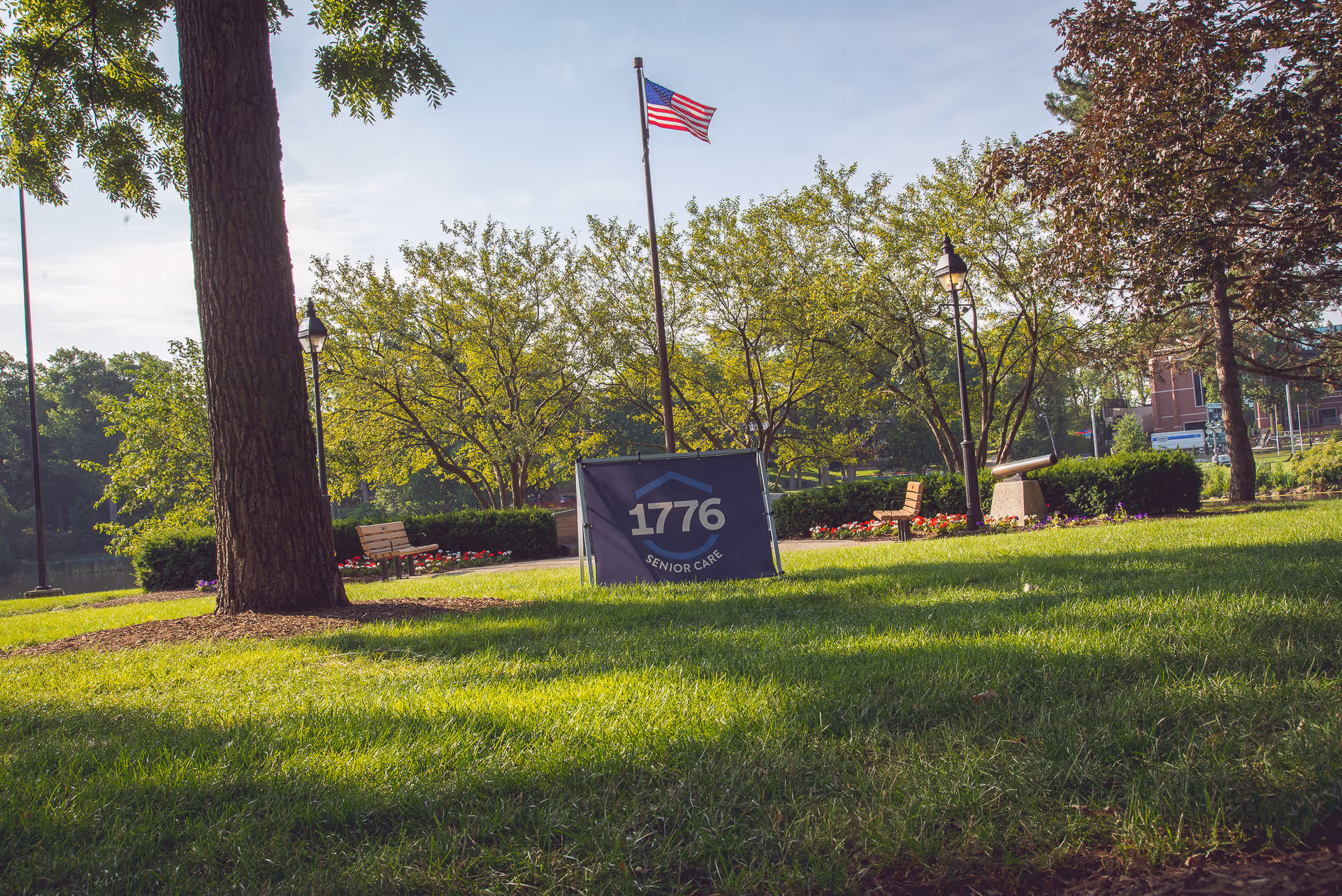 Lush green park with benches, trees, and a 1776 Senior Care sign under an American flag