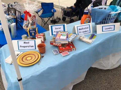 Blue tablecloth booth with labeled activity signs and care items at an outdoor Visiting Angels display.