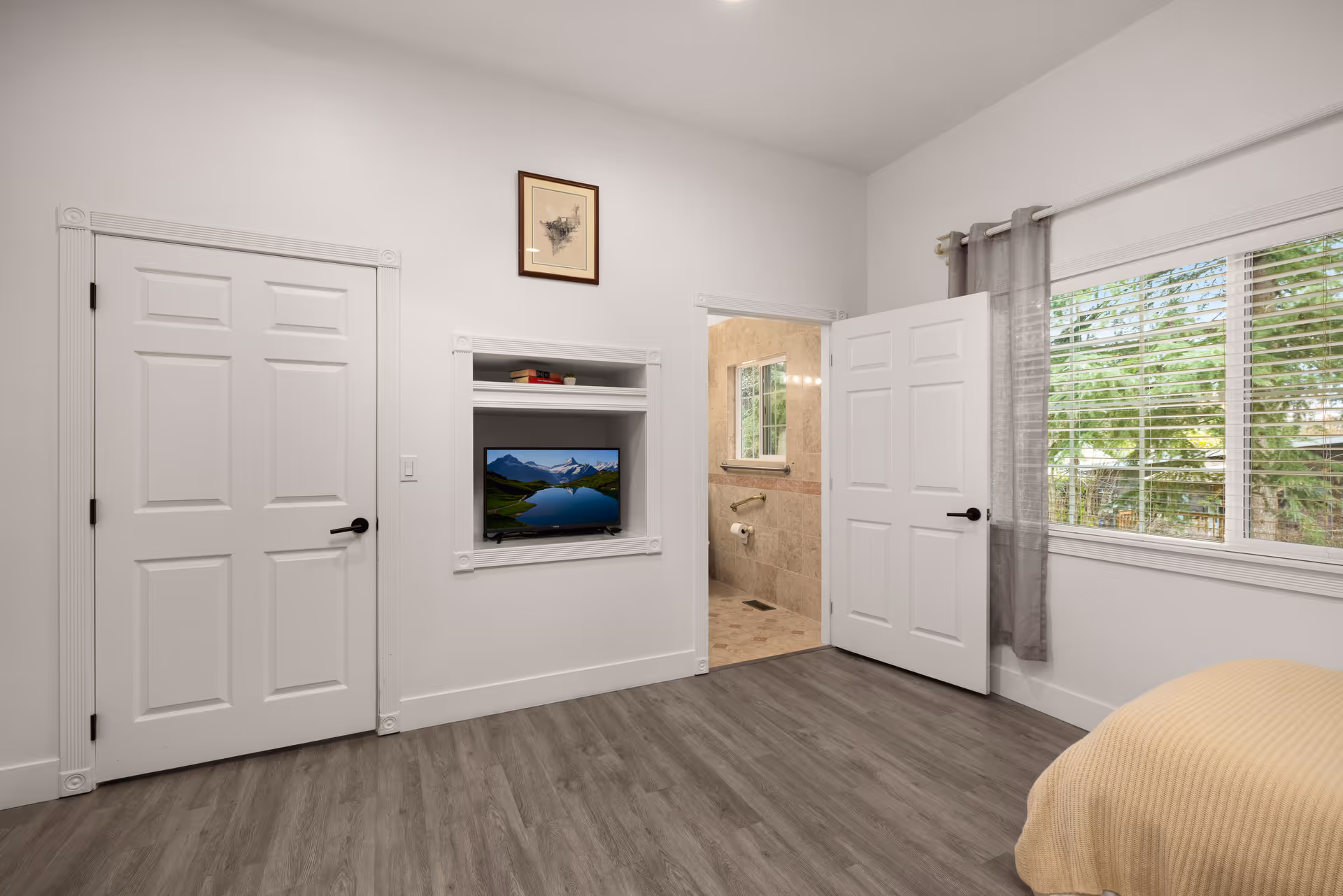 Bright white bedroom with a wall TV nook, two doors, and a large window.