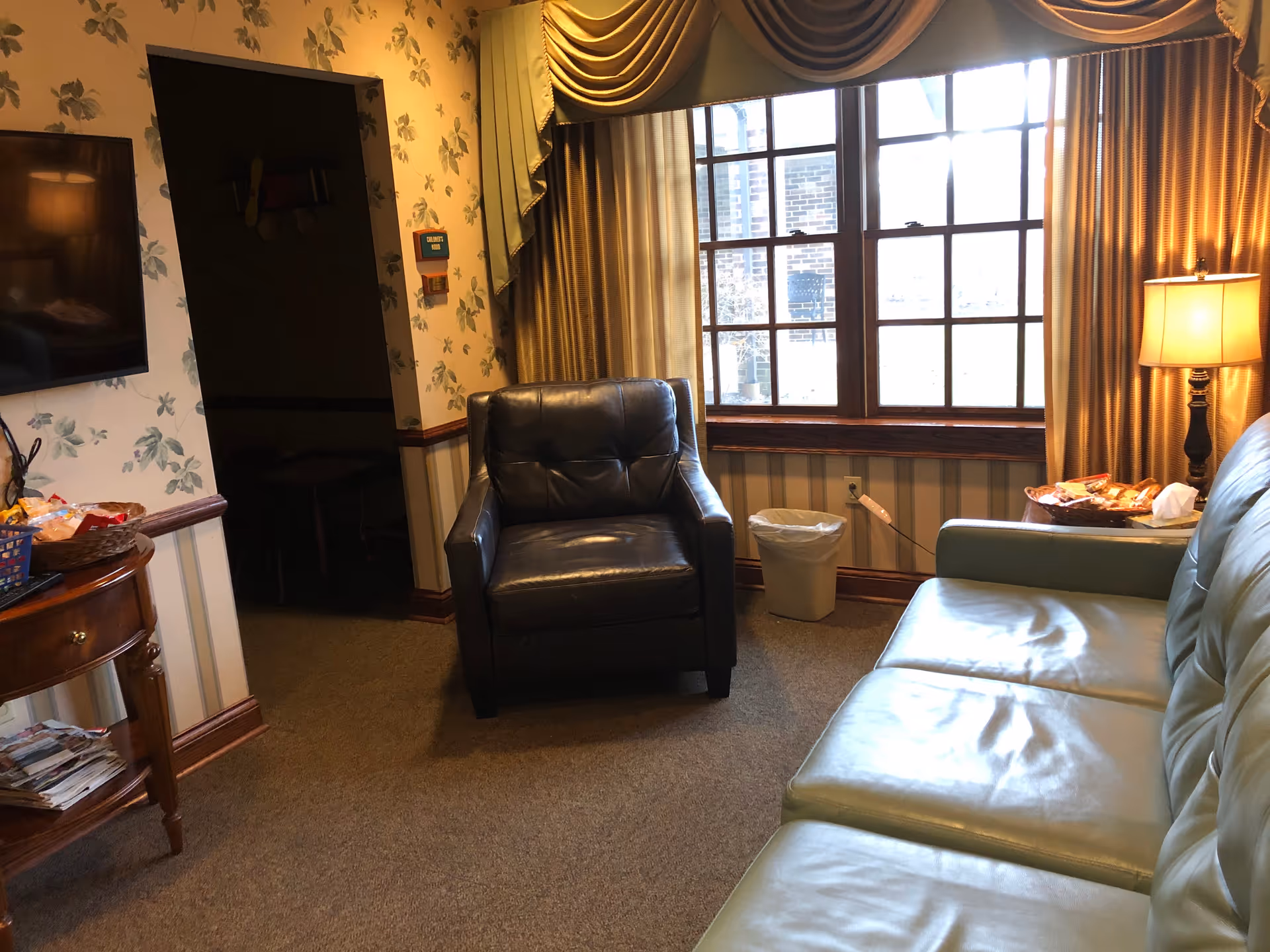A small living room lounge with a leather chair, blue sofa, large window, and warm lighting.