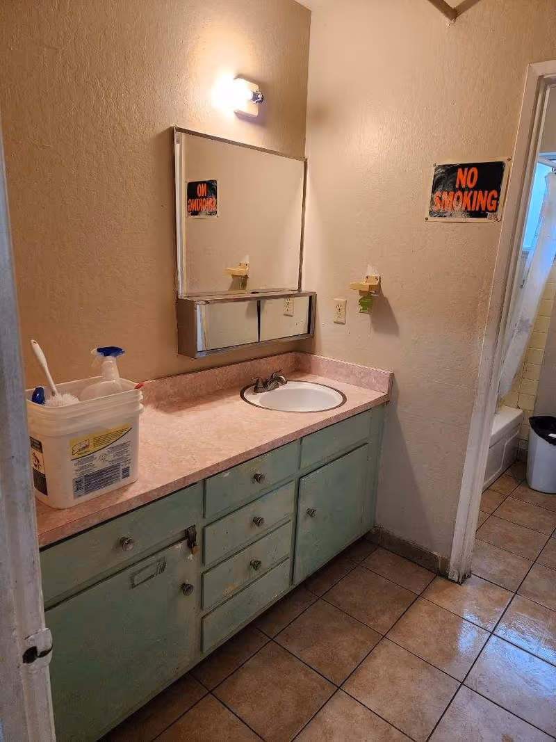A bathroom vanity with mint-green cabinets, pink countertop, sink, mirror, and No Smoking signs.