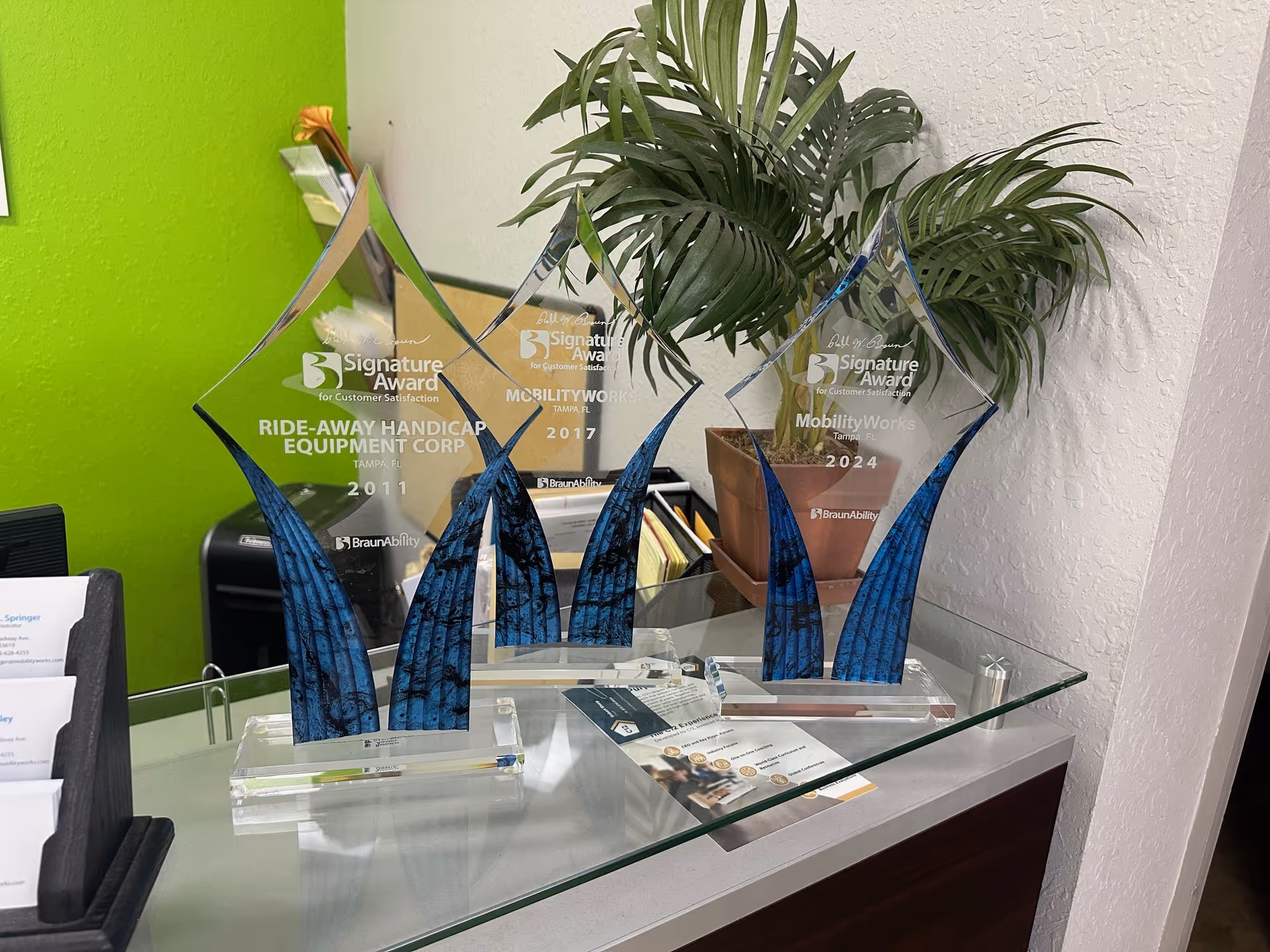 Reception desk with glass awards, green wall, and a plant in a bright lobby.