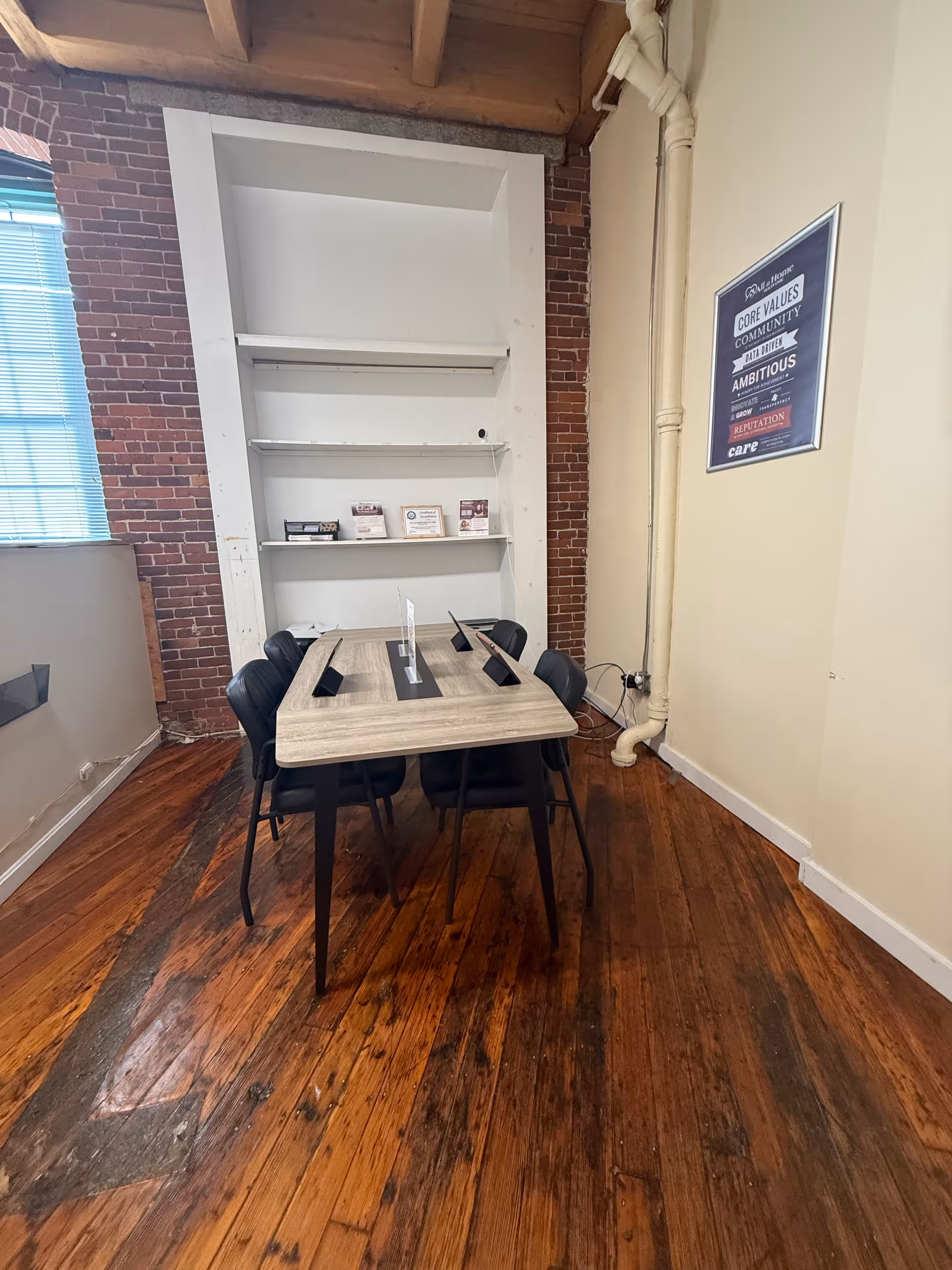 Interior meeting room with a wooden table, chairs, brick wall, and built-in shelves.
