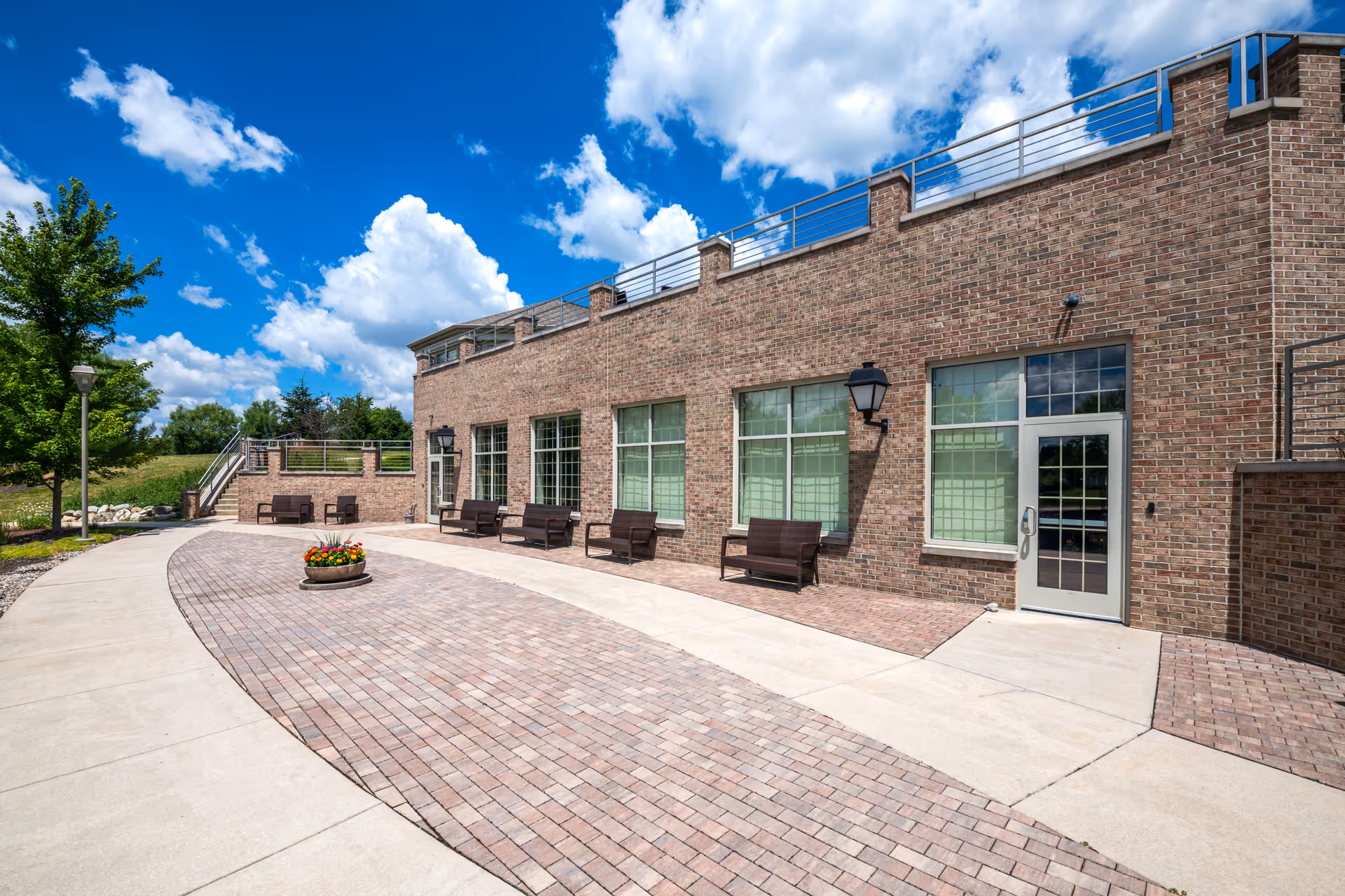 Exterior brick facility with curved brick walkway, outdoor seating benches, and potted flowers.