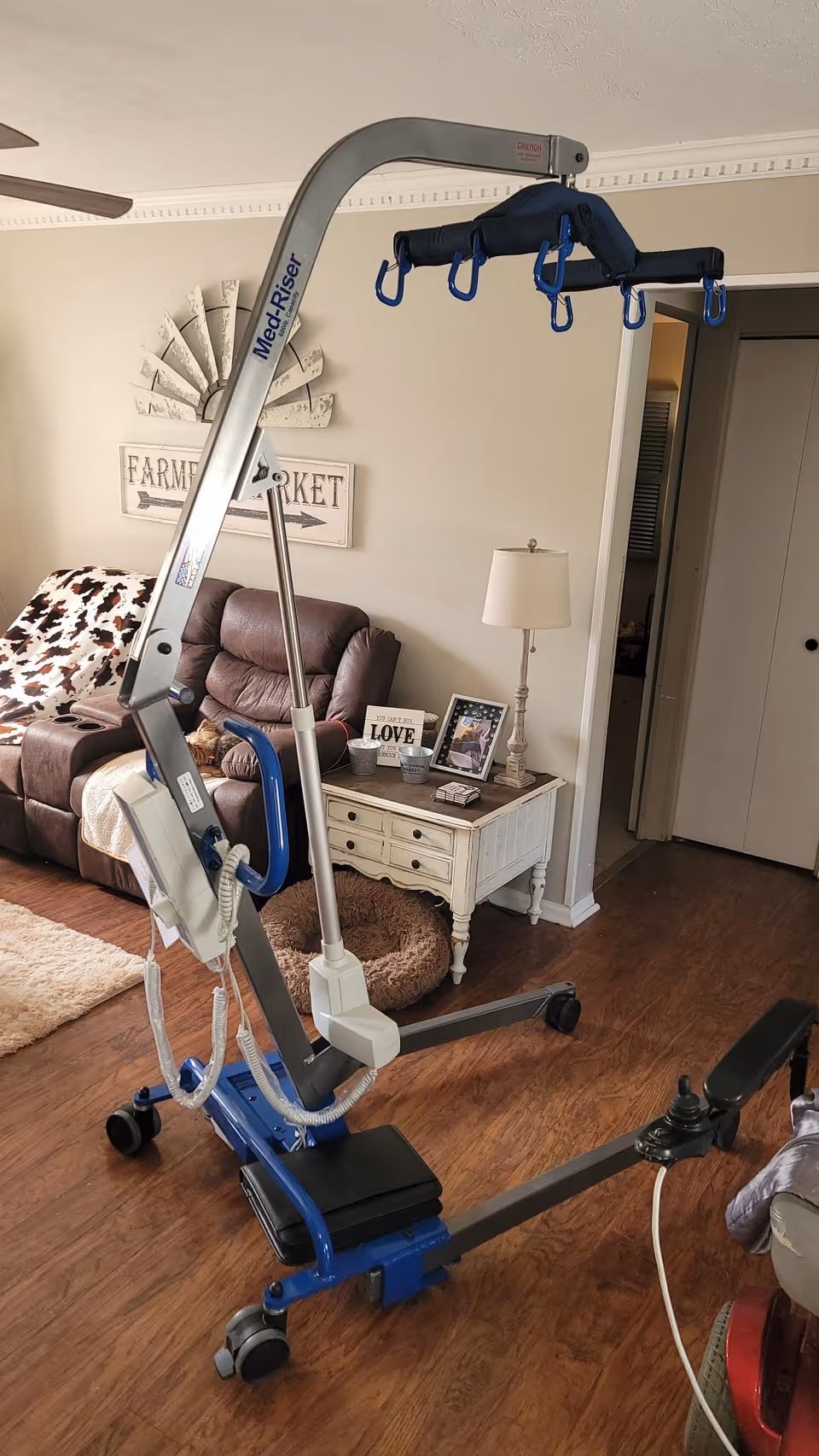 A Mobility Plus patient lift stands in a home living room beside a sofa.