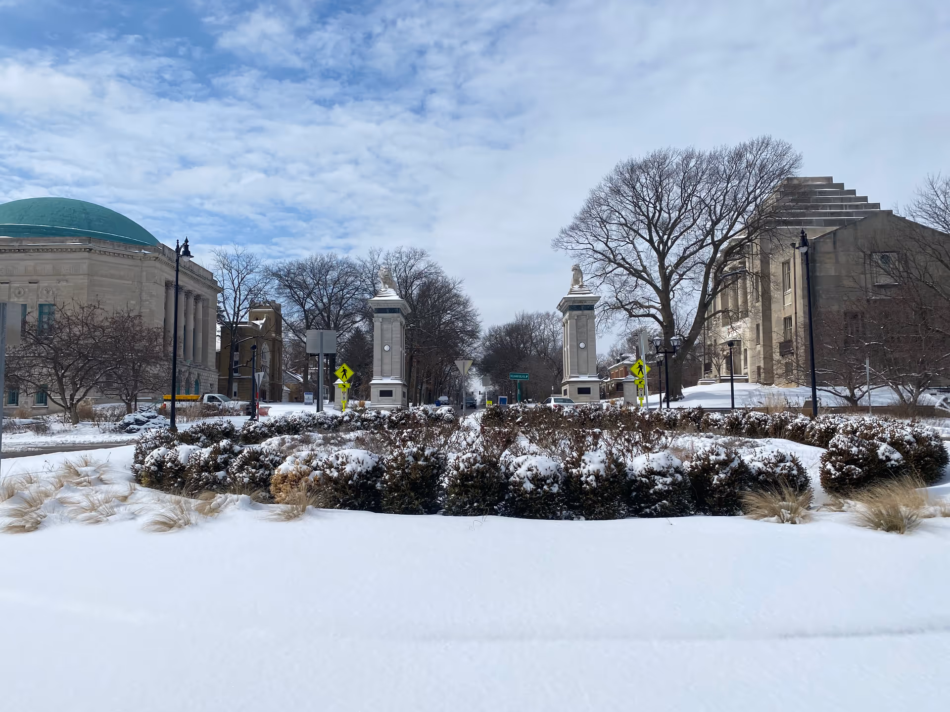 Snow-covered campus street with buildings, trees, and tall stone pillars against a blue sky.
