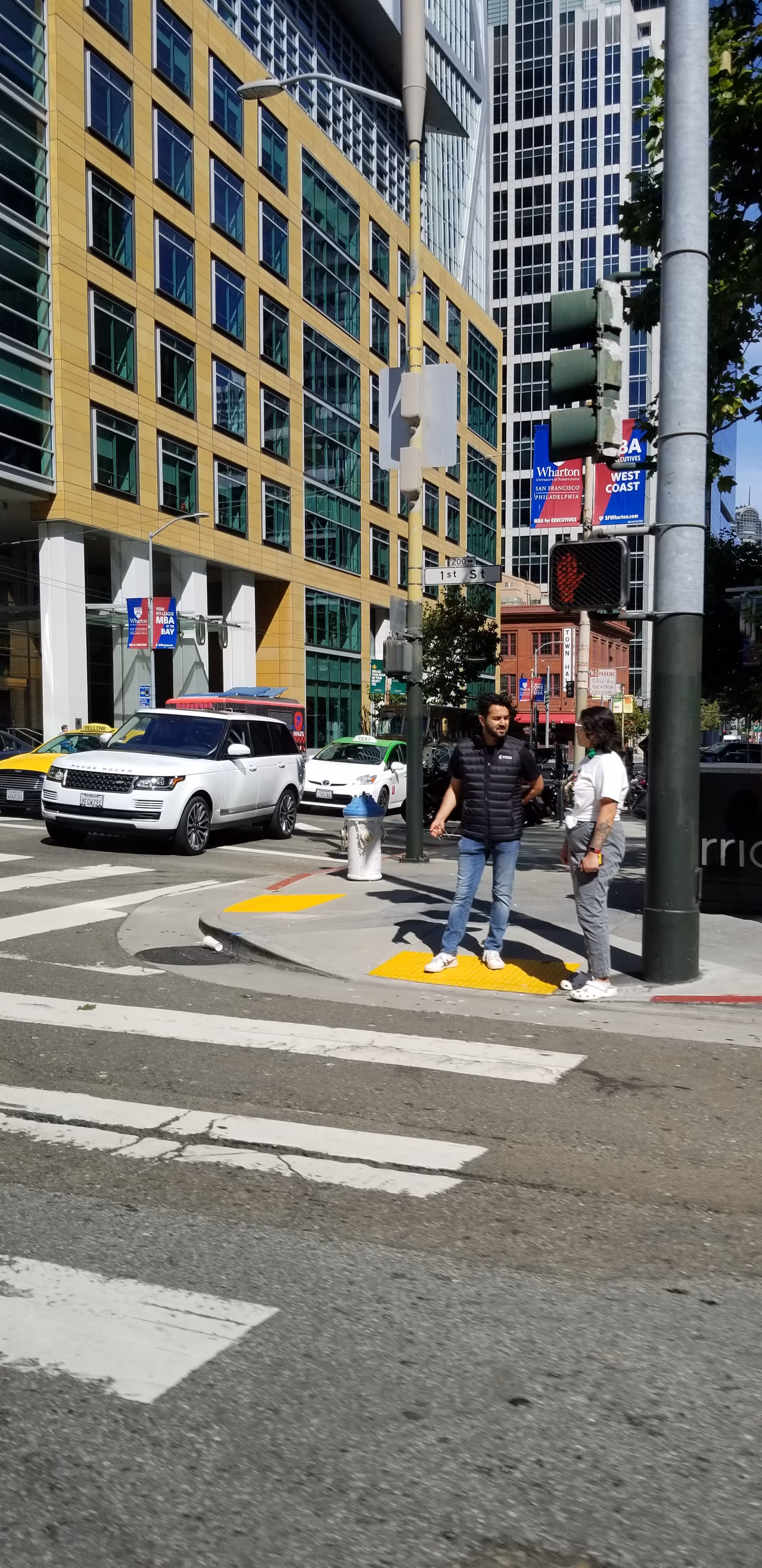 Two people stand on a crosswalk beside cars among tall urban buildings.