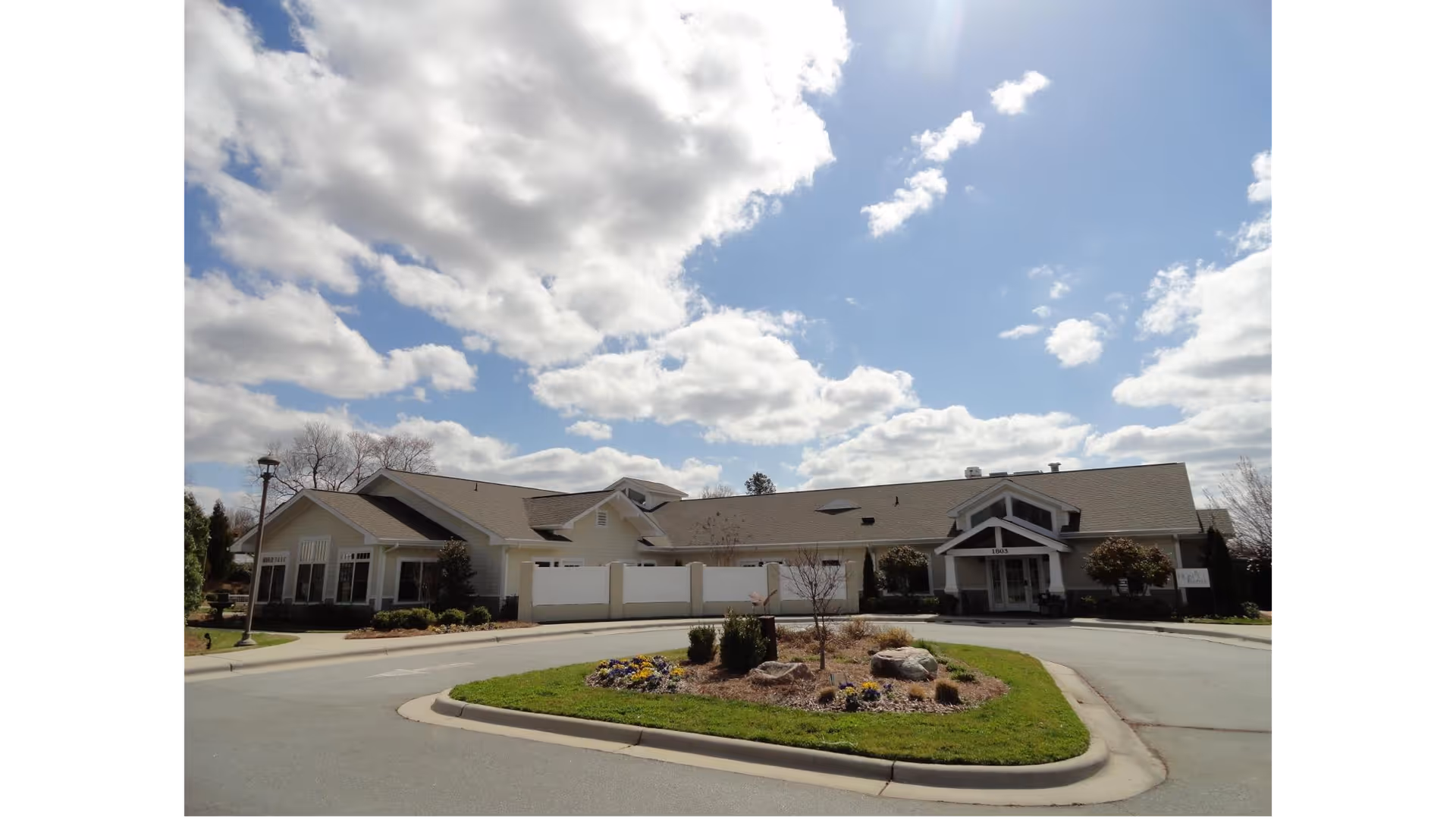 Beige, single-story hospice building with circular driveway and landscaped front, under a cloudy blue sky.