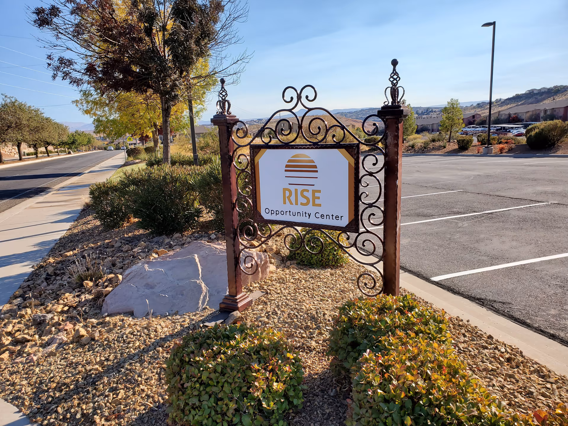 Ornate wrought-iron sign reading RISE Opportunity Center stands among desert landscaping near a parking lot.