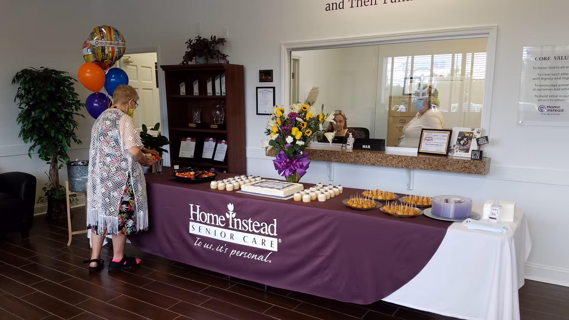 Reception area with a purple Home Instead banner, cupcakes, flowers, and staff behind the counter.