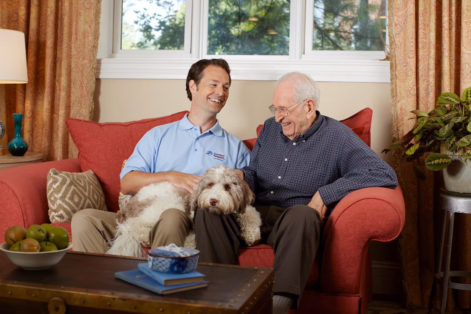 Caregiver and elderly man lounge on a red sofa with a dog.