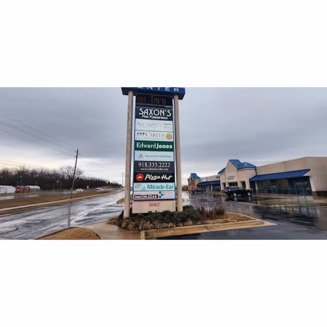 Tall outdoor shopping plaza sign with multiple business listings and a storefront in the distance.