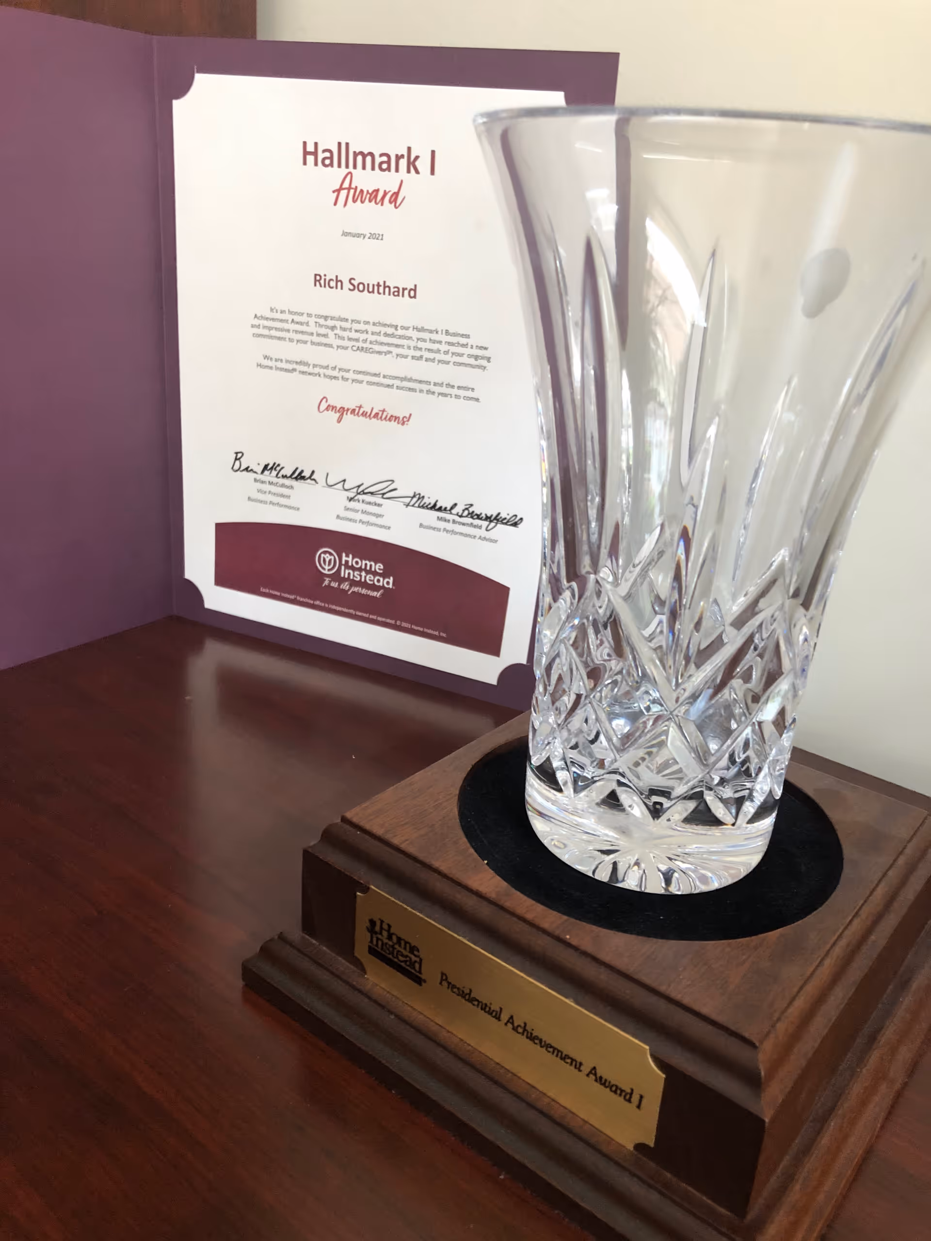 Crystal trophy on wooden desk beside a purple Hallmark Award certificate.