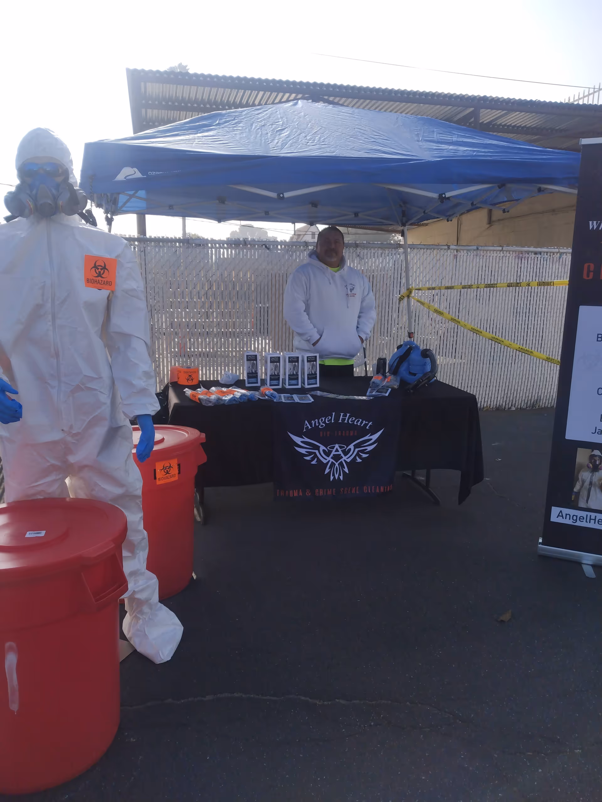 Two people operate an outdoor health booth under a blue canopy displaying PPE gear.