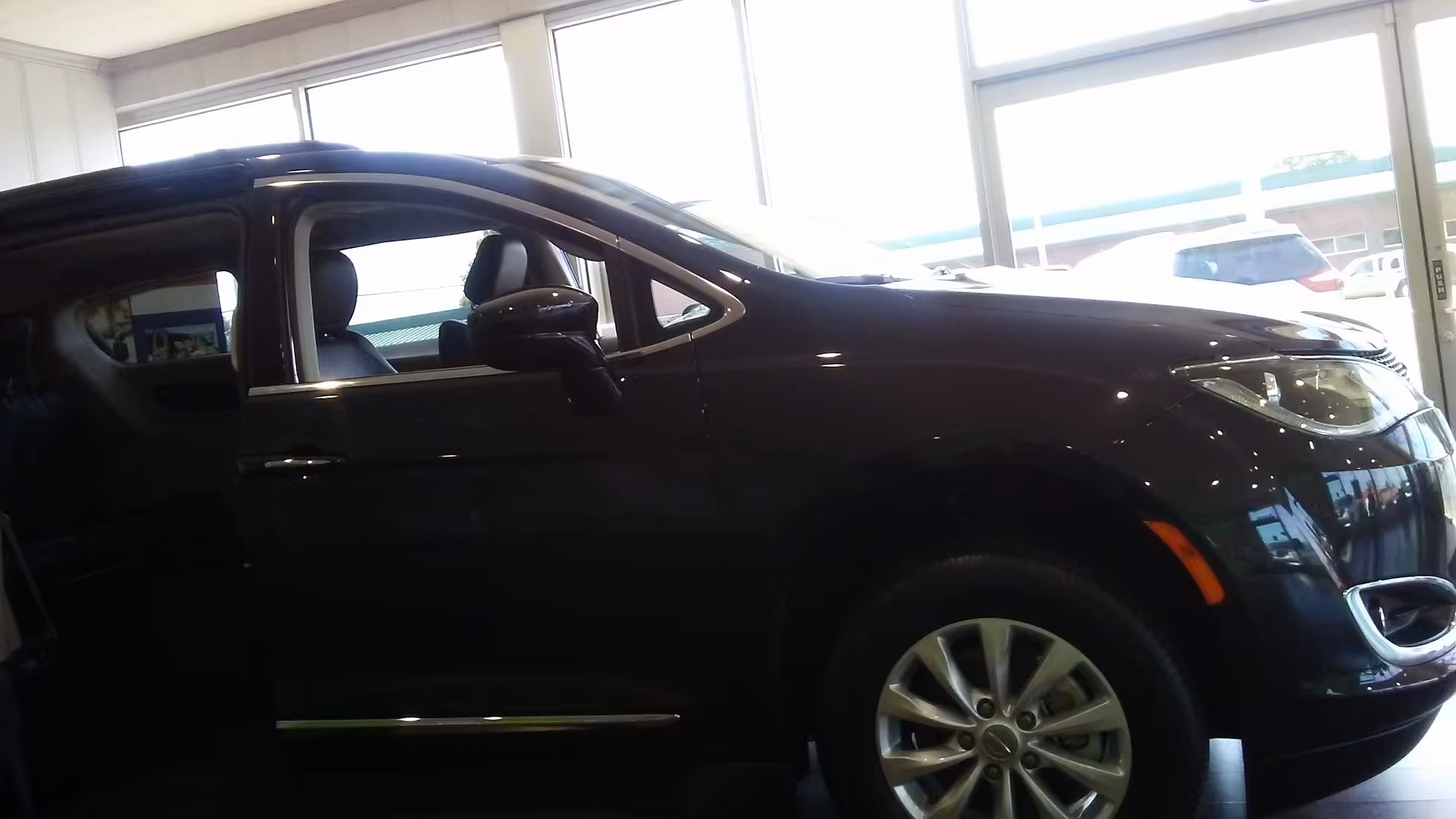 A dark car is parked indoors in a showroom with large windows and outside vehicles.