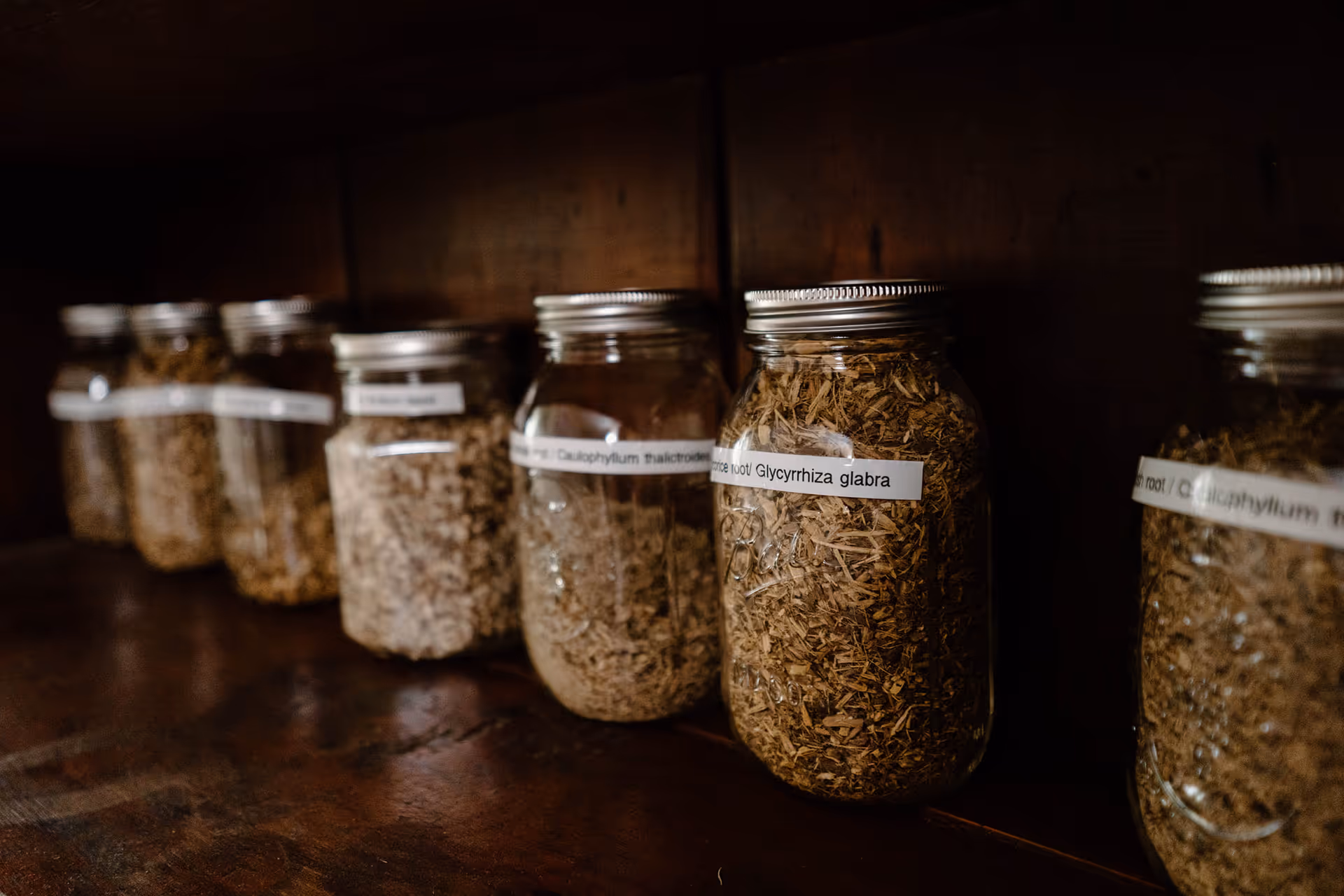 Row of glass jars with labeled dried herbs on a wooden shelf.