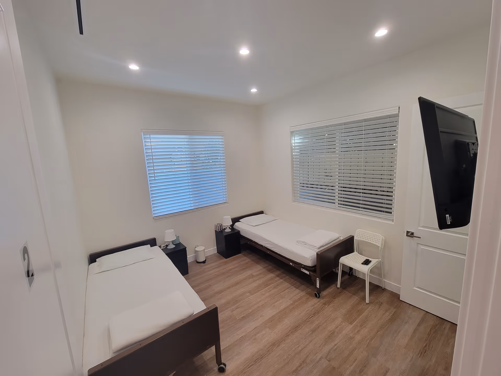 Two single beds in a small white bedroom with blinds, nightstands, and a wall-mounted TV.