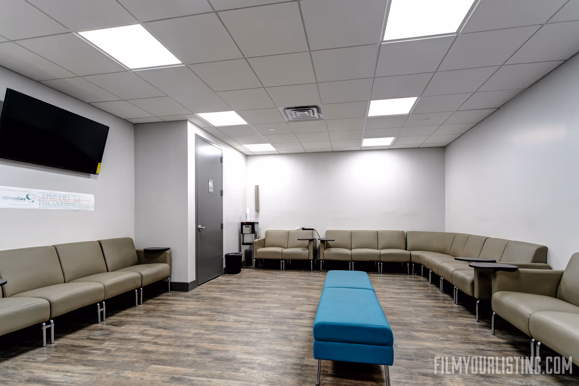 Bright waiting area with beige modular sofas, a turquoise ottoman, and a wall-mounted TV.