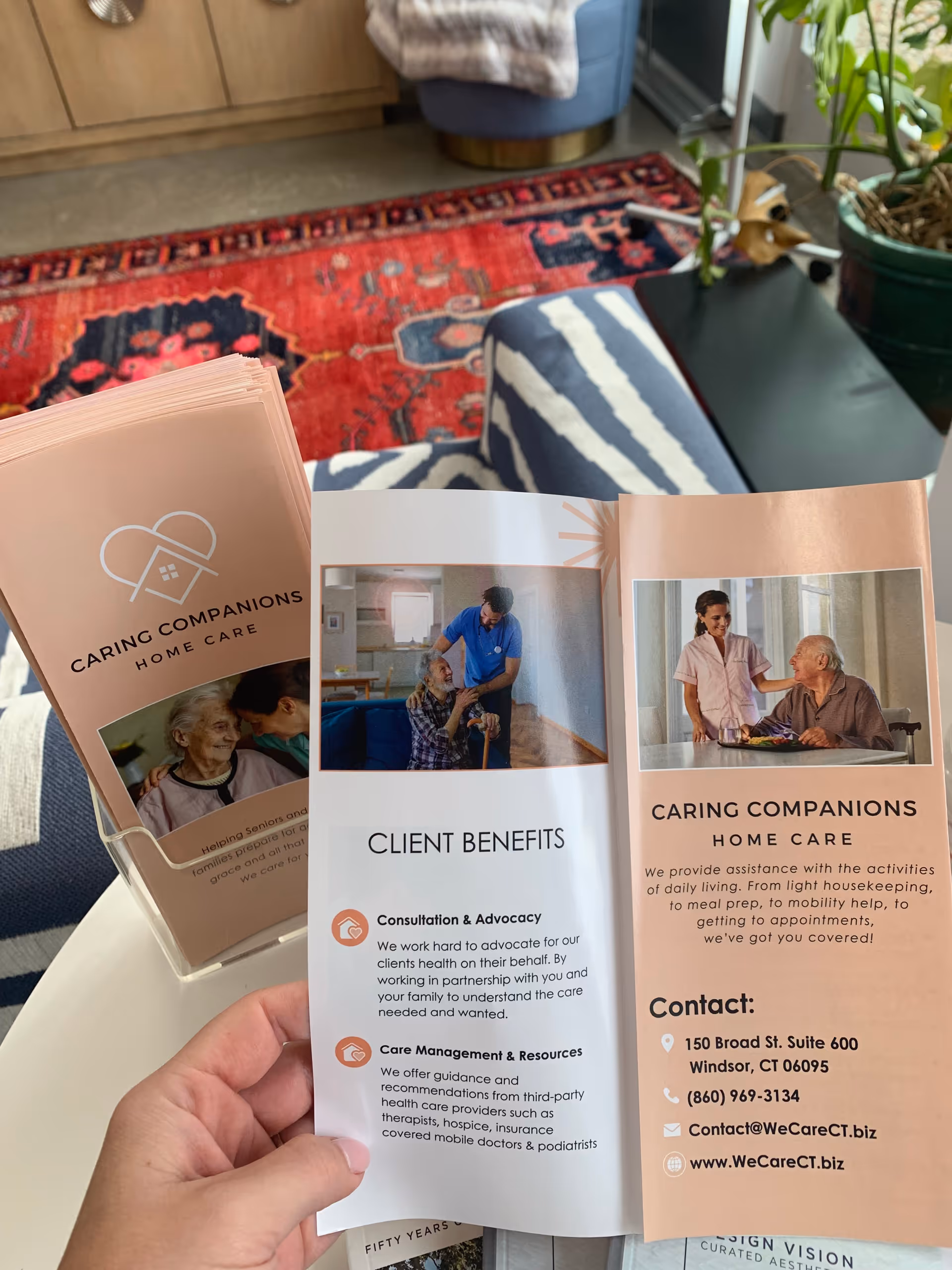 Hand holds Caring Companions Home Care brochures in a cozy living room.