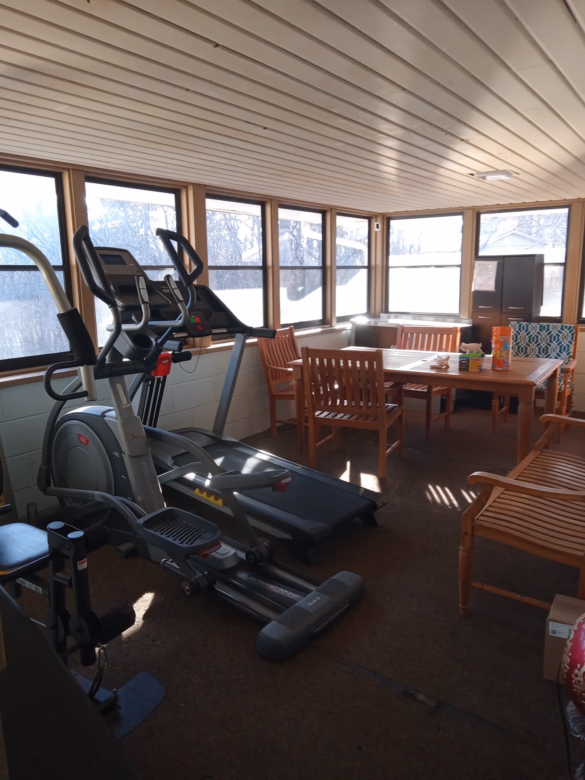 Sunlit indoor common area with treadmills and a wooden dining table for residents.