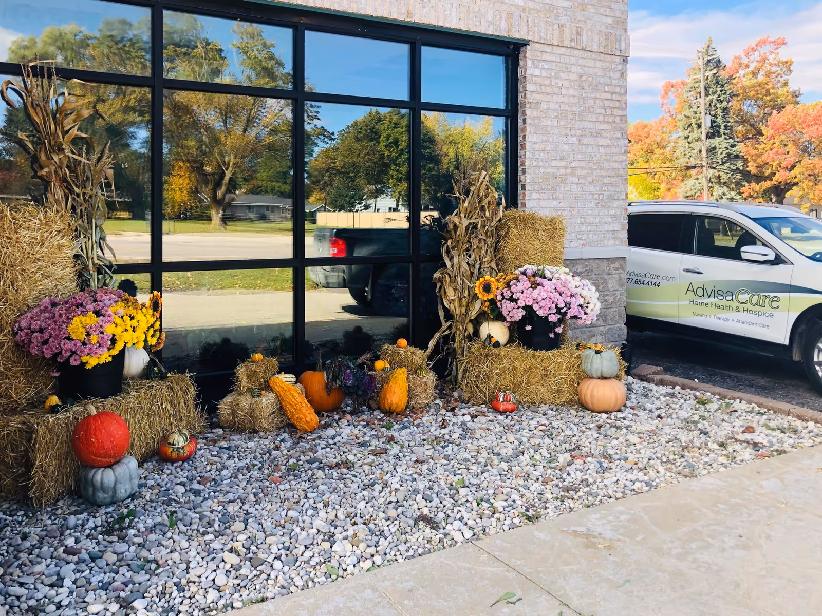 Fall decorations with pumpkins, hay bales, and flowers decorate building front beside a branded AdviaCare van.