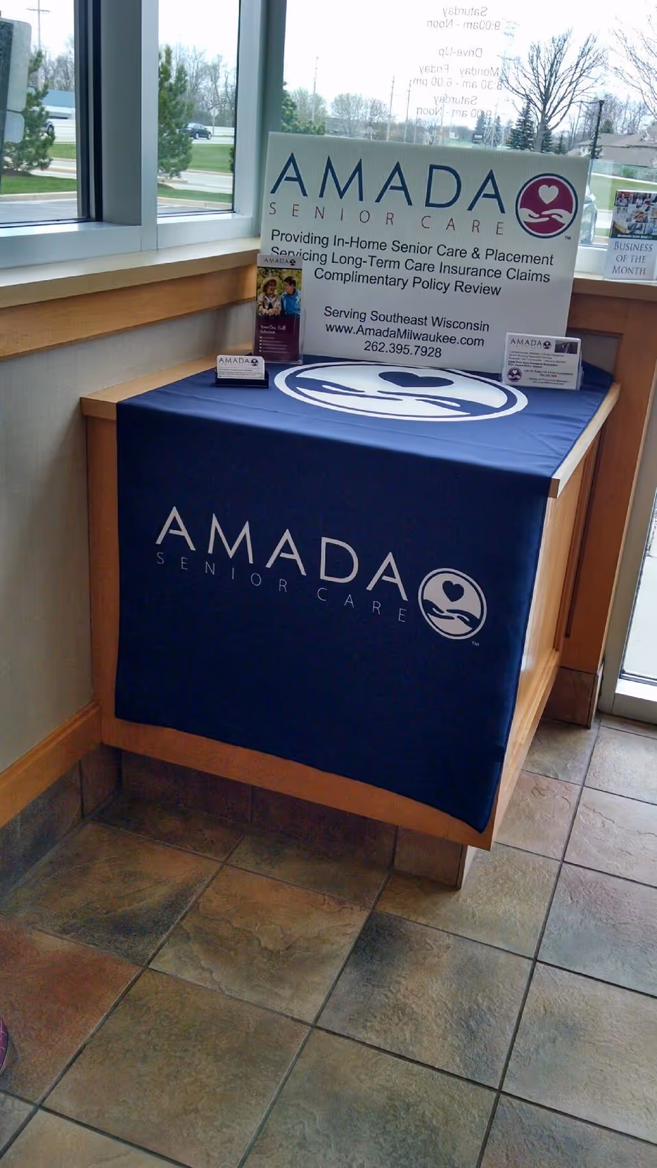 Amada Senior Care display sign on a blue table in a lobby near windows.