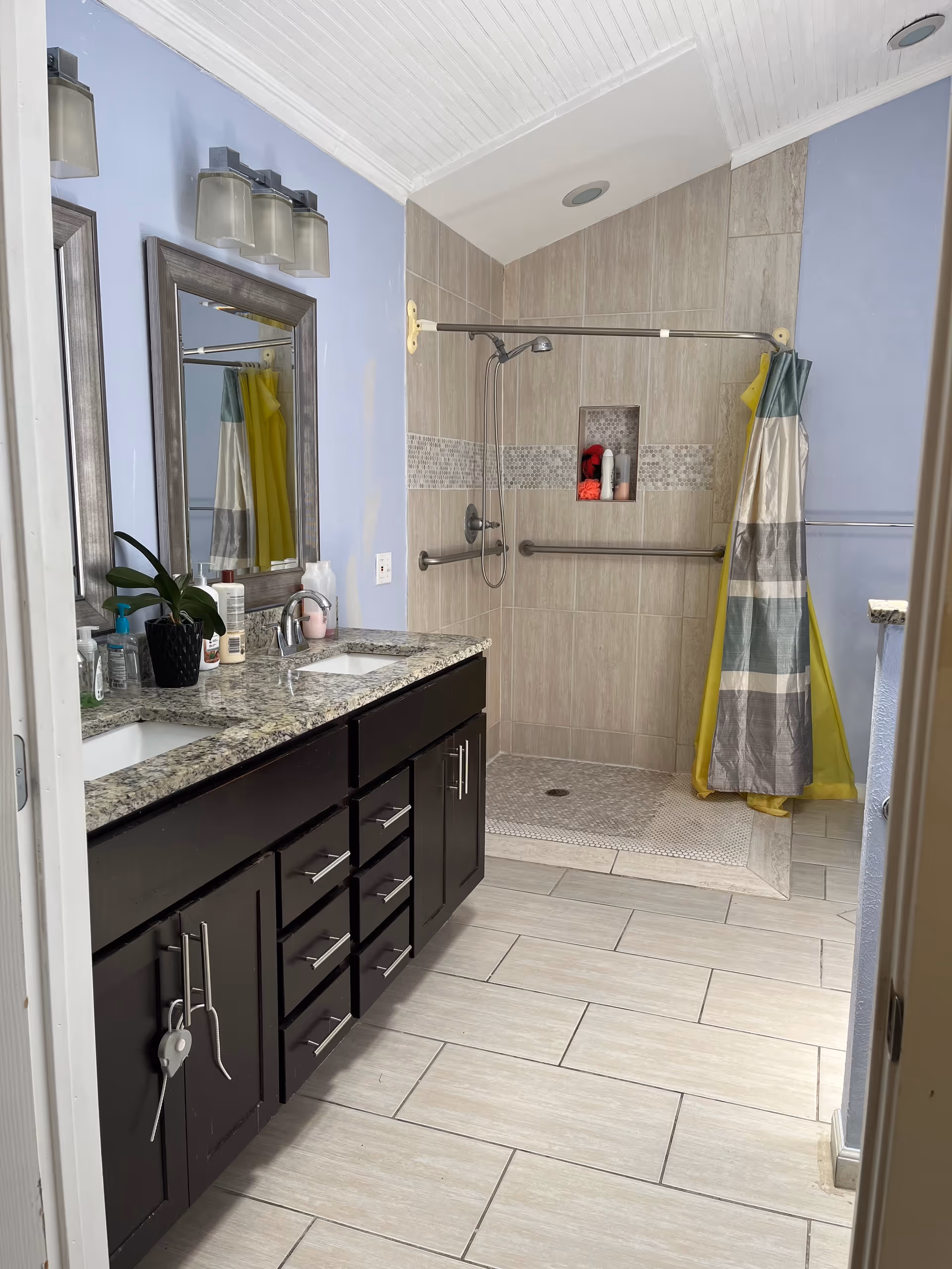 A contemporary bathroom with double vanity, granite countertop, large mirror, and accessible shower.