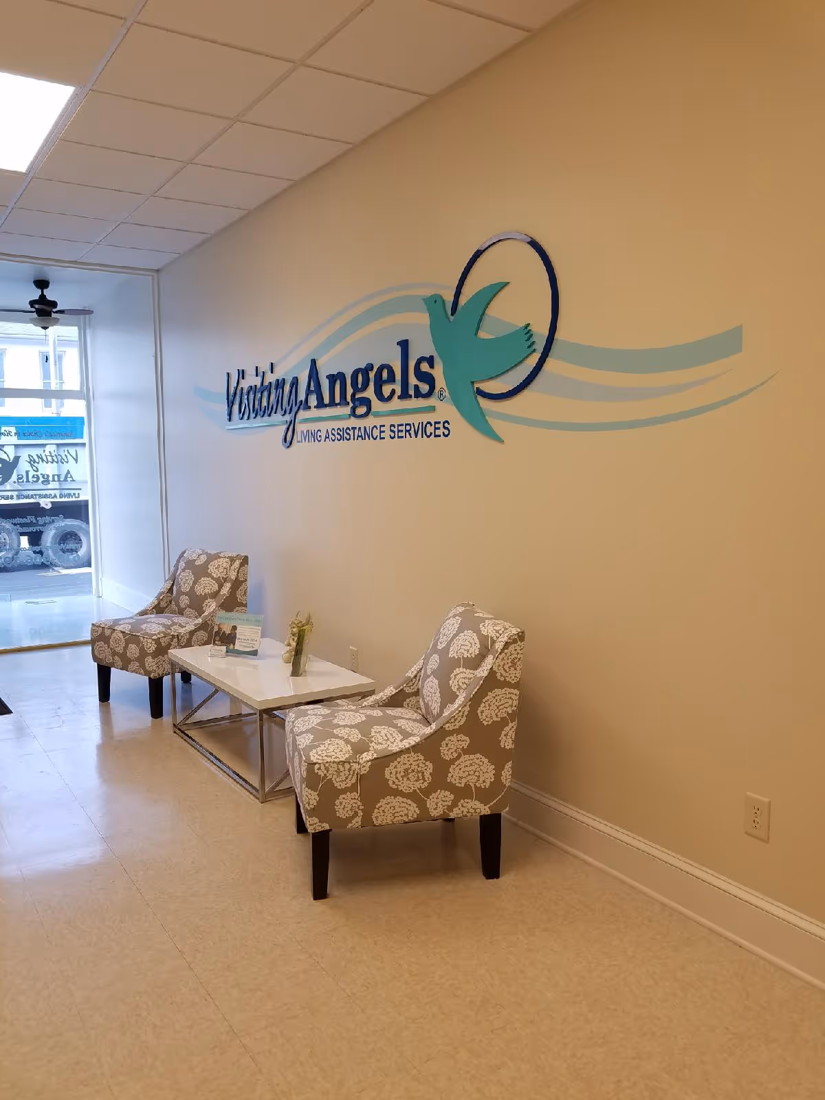 Lobby seating area with two floral chairs, a glass table, and a wall logo Visiting Angels.