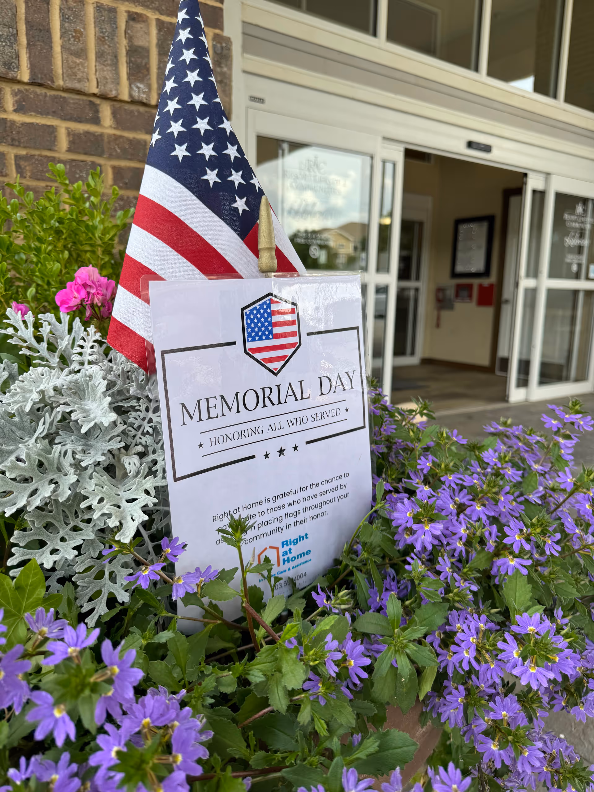 Memorial Day display with American flag outside Right at Home entrance amid purple flowers.