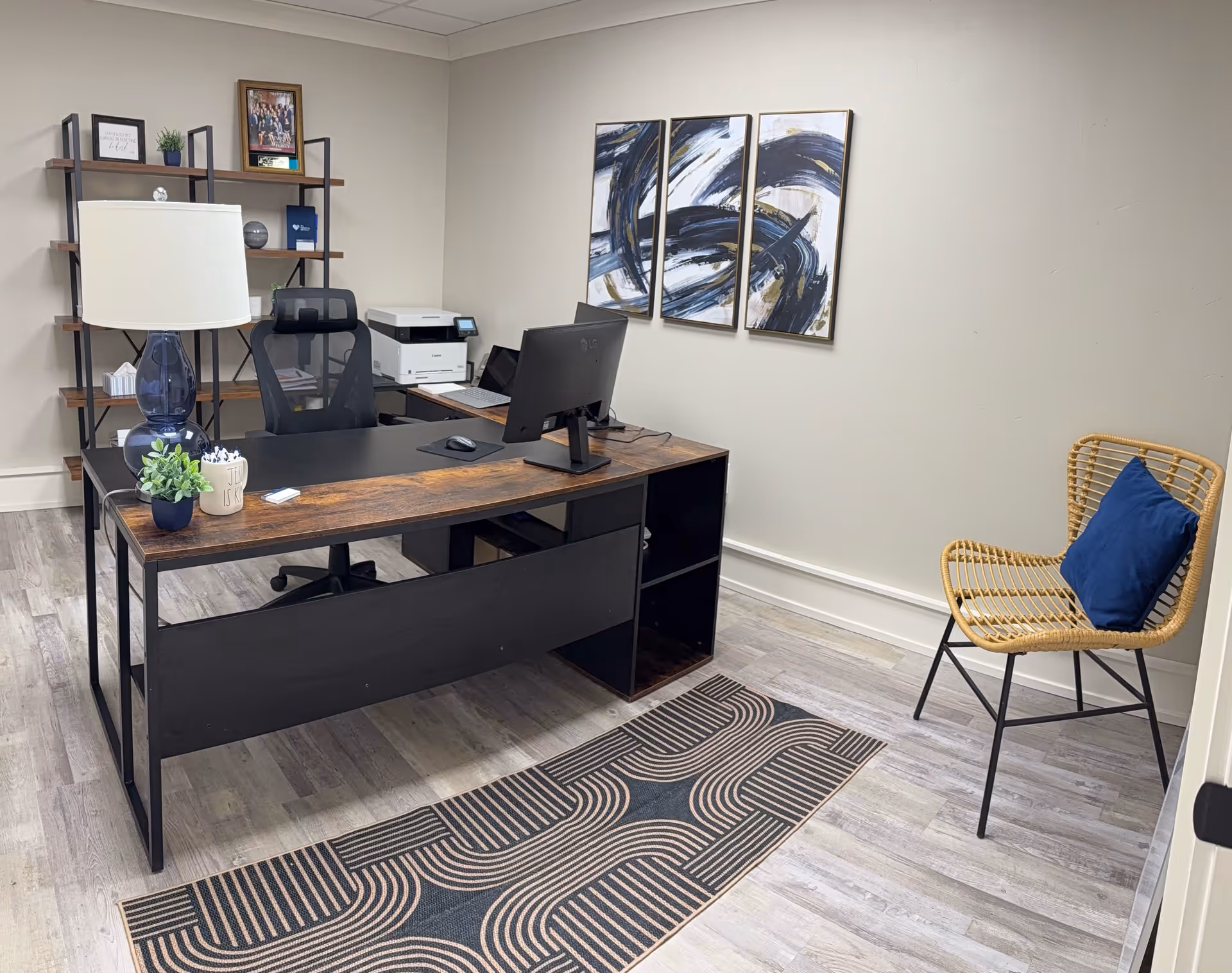 Modern office desk setup with computer, printer, lamp, shelves, and a wicker chair.