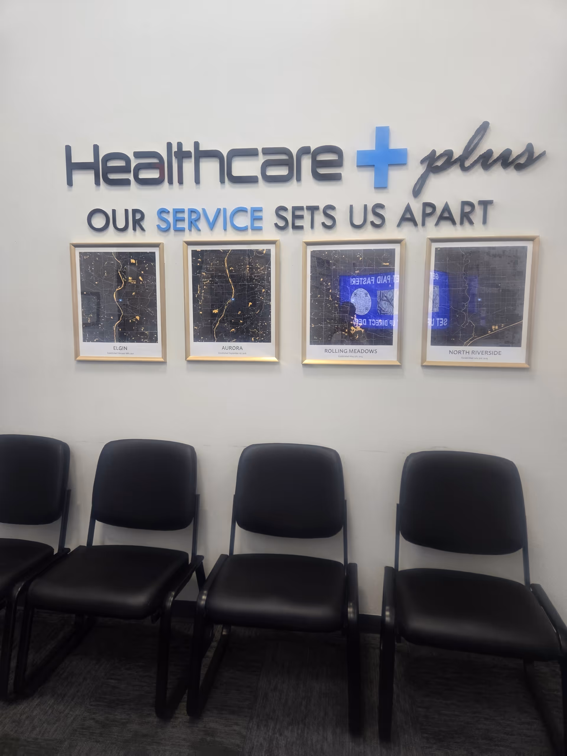 Interior lobby area with Healthcare Plus sign, slogan, framed maps, and black waiting chairs.