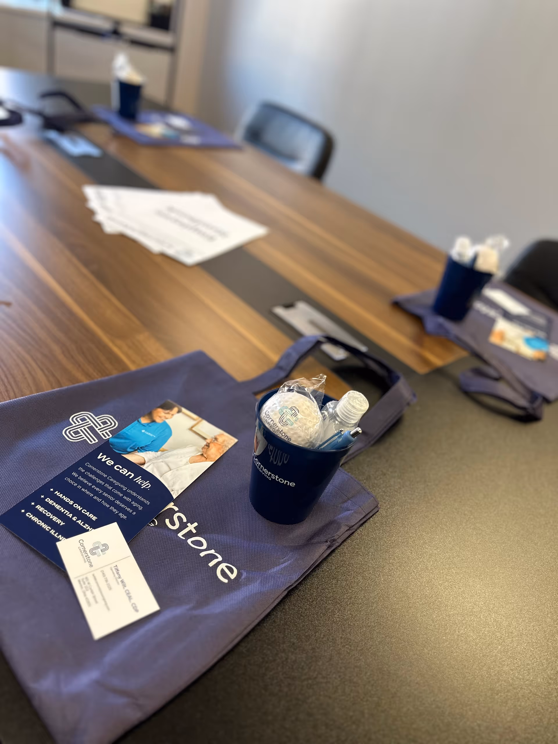 A conference room table with a Cornerstone Caregiving branded bag, pamphlets, and a blue cup.
