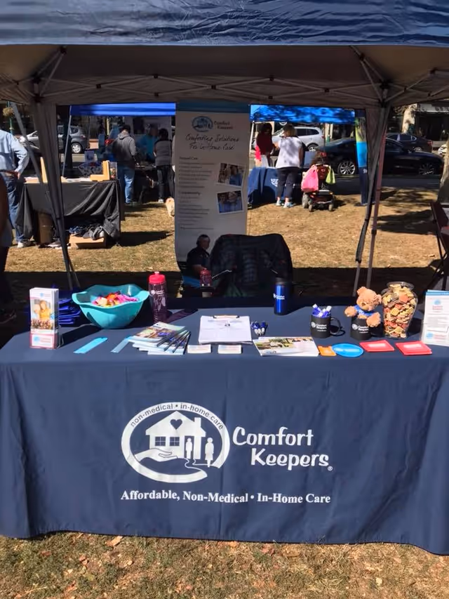 Outdoor Comfort Keepers booth with banner, tables, and attendees under canopies.